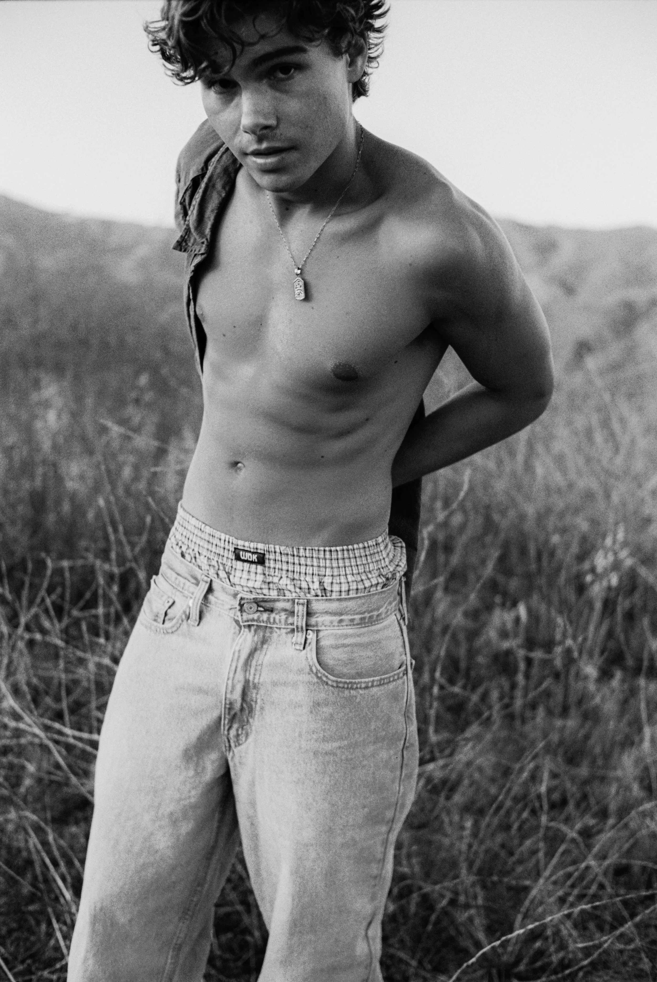 A young man with curly hair posing outdoors shirtless, wearing jeans and a dog tag necklace, with mountains and tall grass in the background.