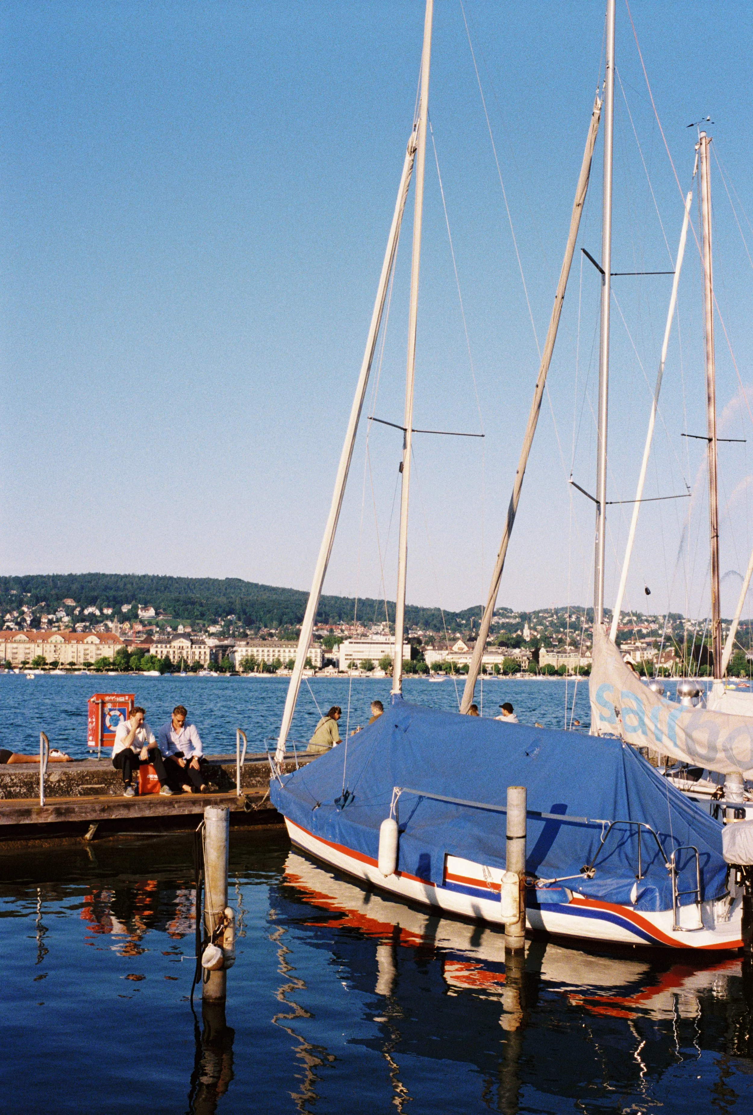 People sitting on a dock near sailboats moored in a harbor with city buildings and green hills in the background.