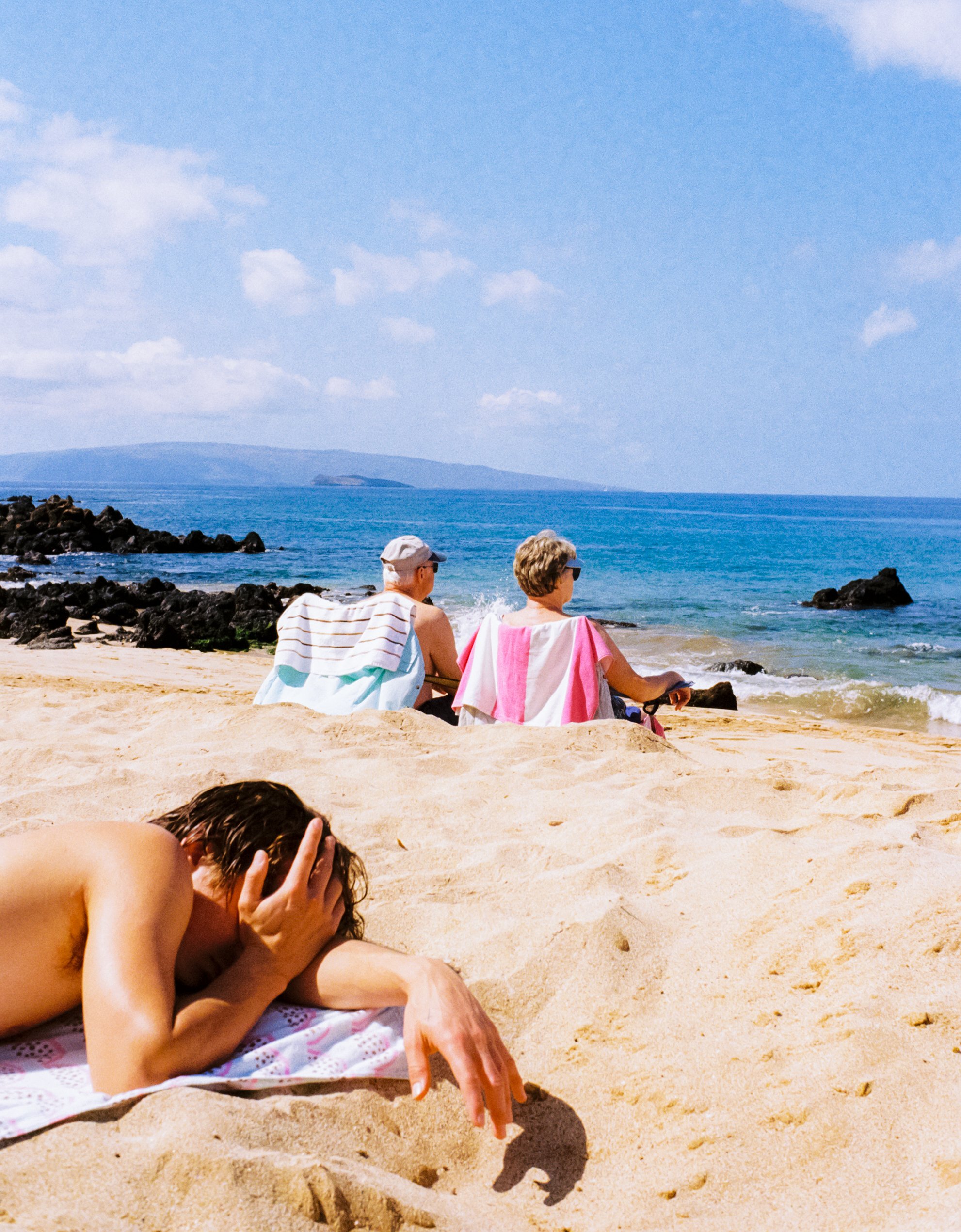 Two elderly women sitting on beach chairs near the ocean, with rocks and a distant island in the background. A young person is lying on sand in the foreground, covering their face with hands.