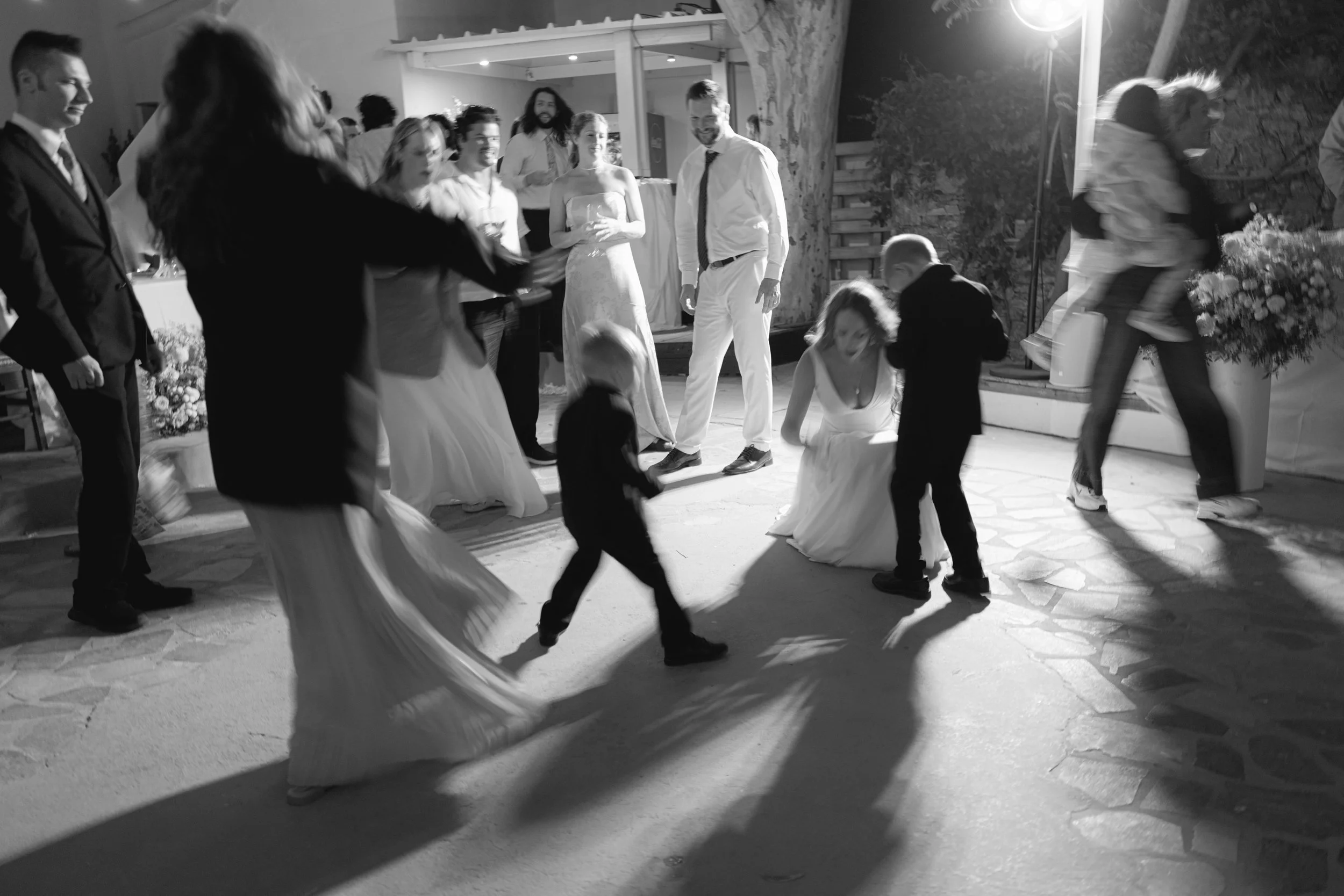 People dancing and socializing at an outdoor evening event, with some women in long dresses and men in formal attire, under string lights near a rustic wooden wall.