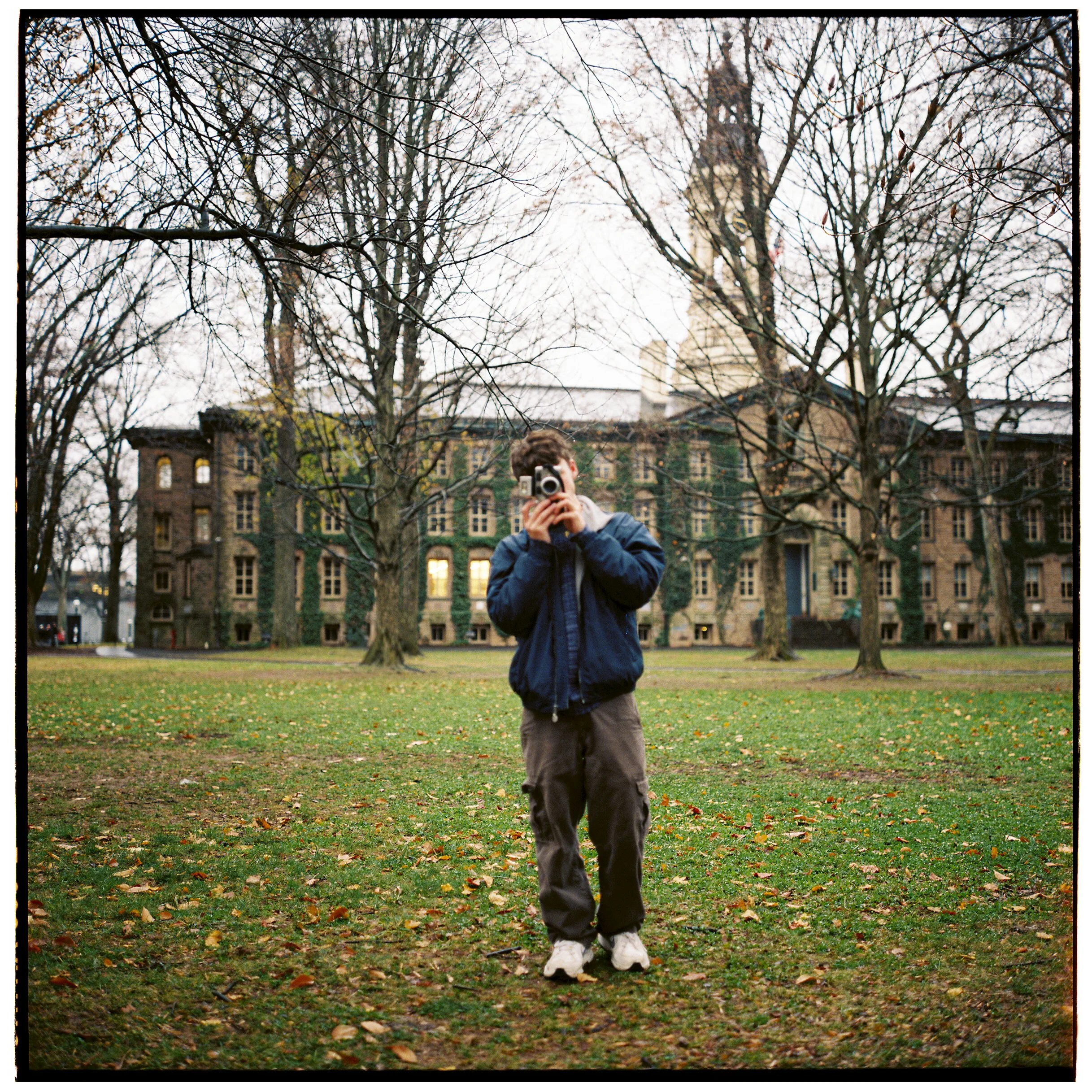 Person standing in a park with trees and fallen leaves, holding a camera up to their face. In the background, there are buildings and a church steeple.