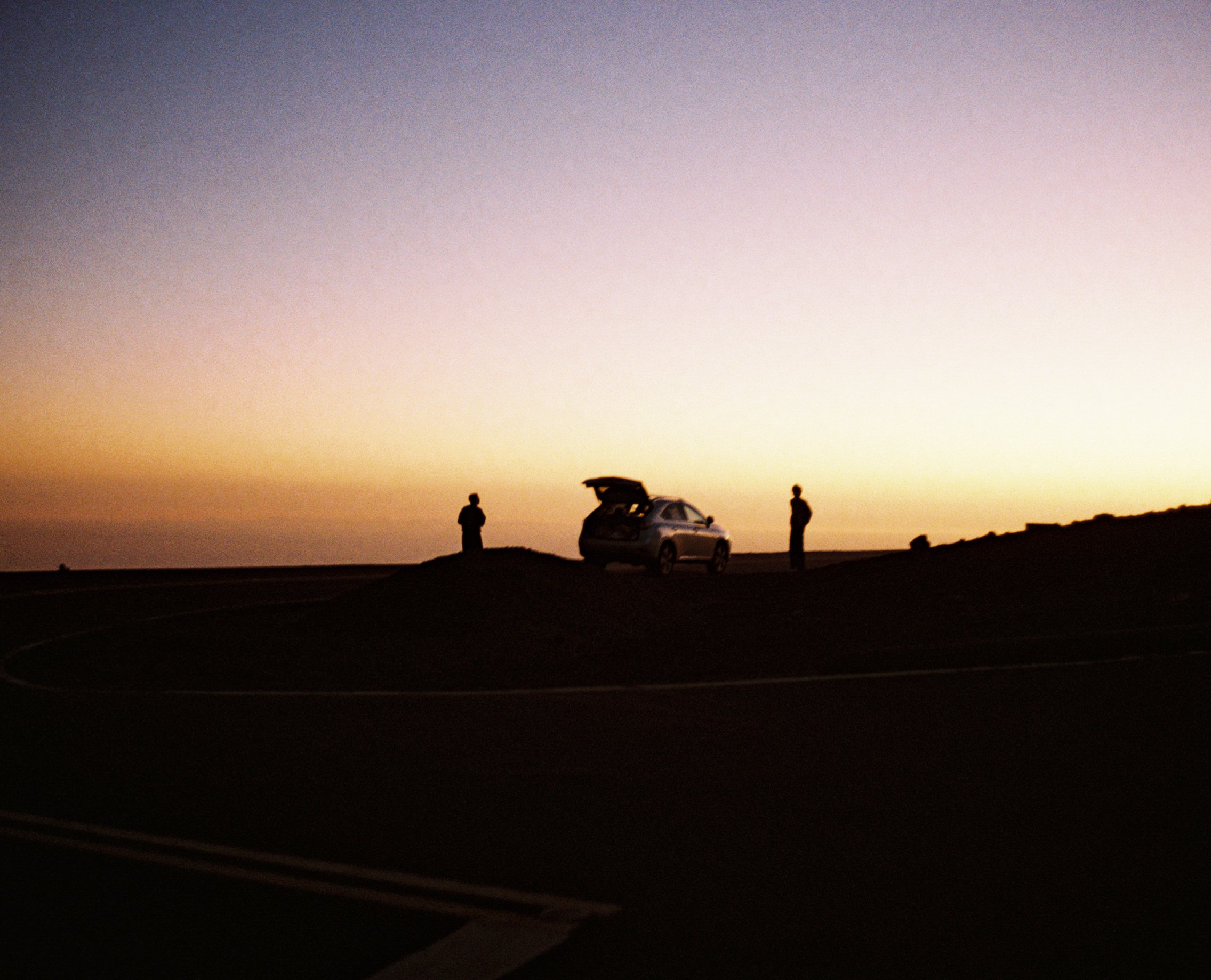 Silhouettes of two people standing near an open car trunk on a hilltop at sunset, with a colorful sky in the background.