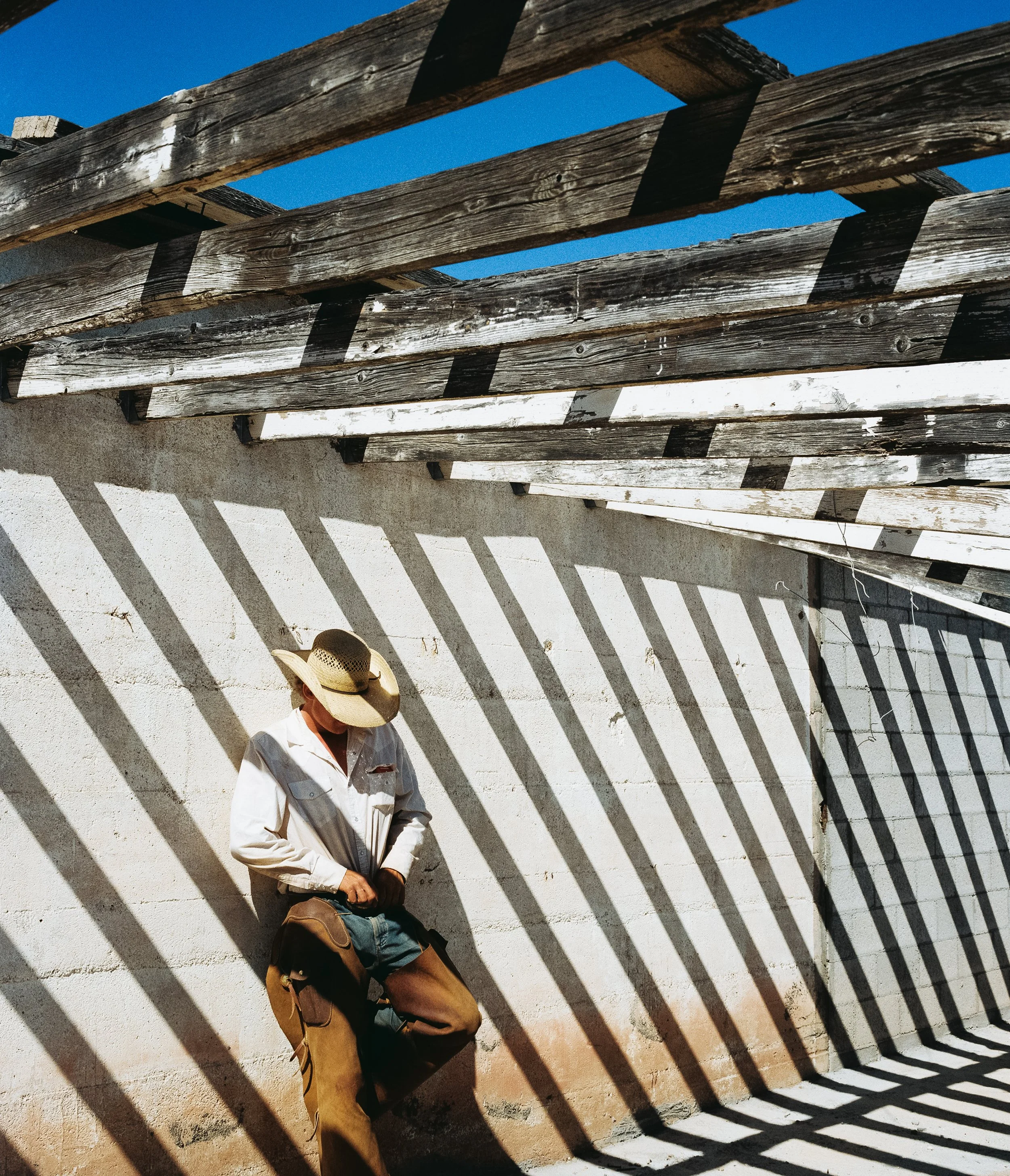 A man wearing a white shirt, tan pants, and a wide-brimmed hat leaning against a white wall with shadow patterns created by wooden beams above, under a clear blue sky.