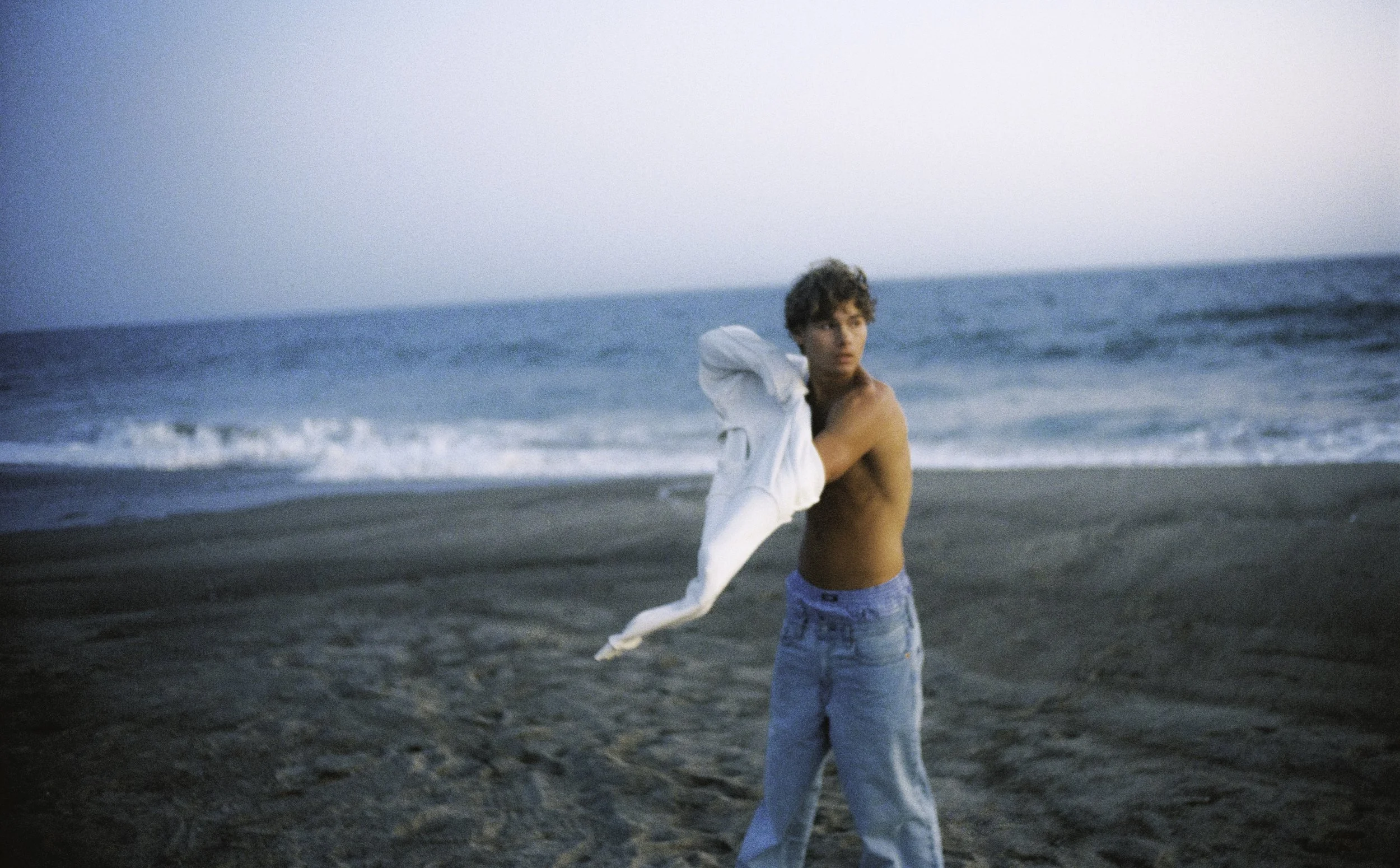 A young man with curly hair, shirtless, wearing light blue jeans, standing on a sandy beach near the ocean, holding a white shirt over his shoulder, looking to the side.