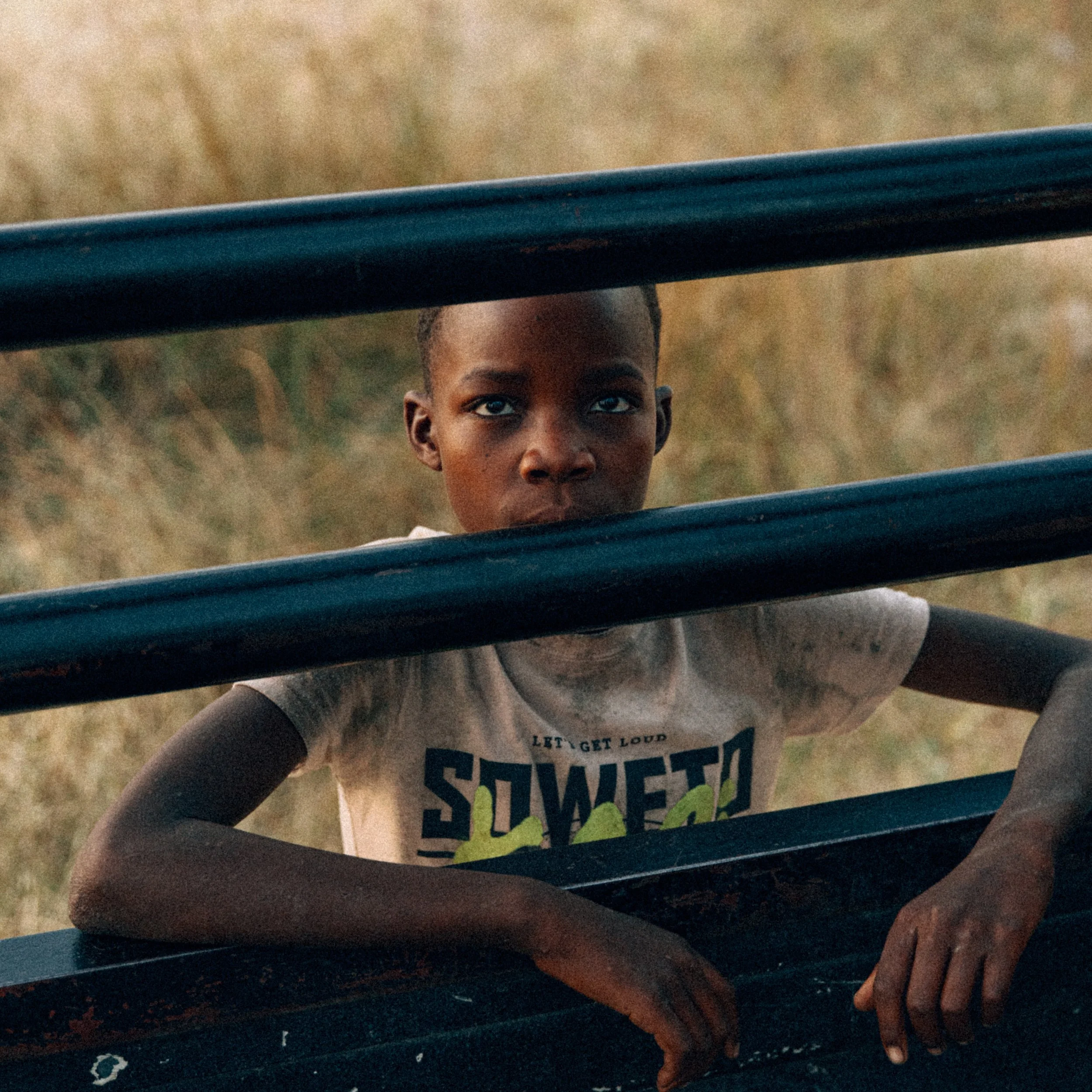 A young boy with dark skin and short hair looking through two horizontal black bars outdoors with a grassy field in the background, wearing a light-colored shirt with the word 'SWEET' printed on it.
