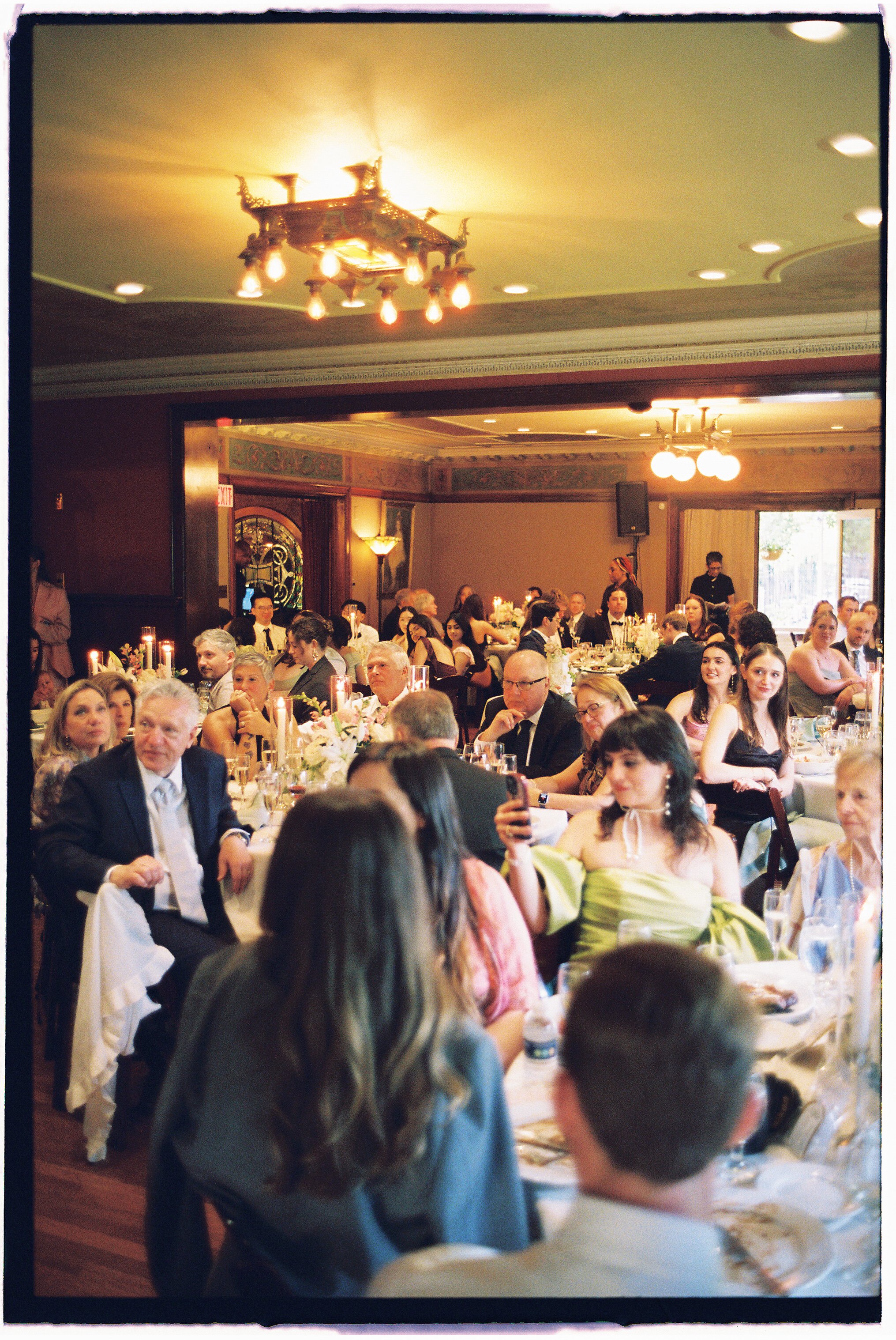 Elegant banquet hall filled with guests seated at decorated tables, with a chandelier hanging from the ceiling, during a formal celebration or wedding reception.
