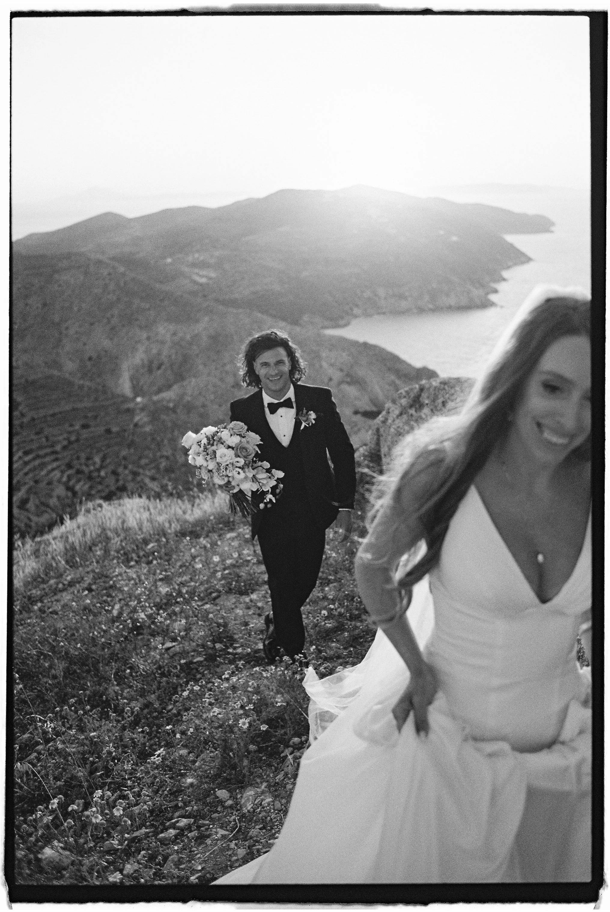 A bride in a white wedding dress with a long train, smiling, standing outdoors on a hilly landscape with mountains and a body of water in the background. A groom in a tuxedo holding a bouquet of flowers is seen behind her.