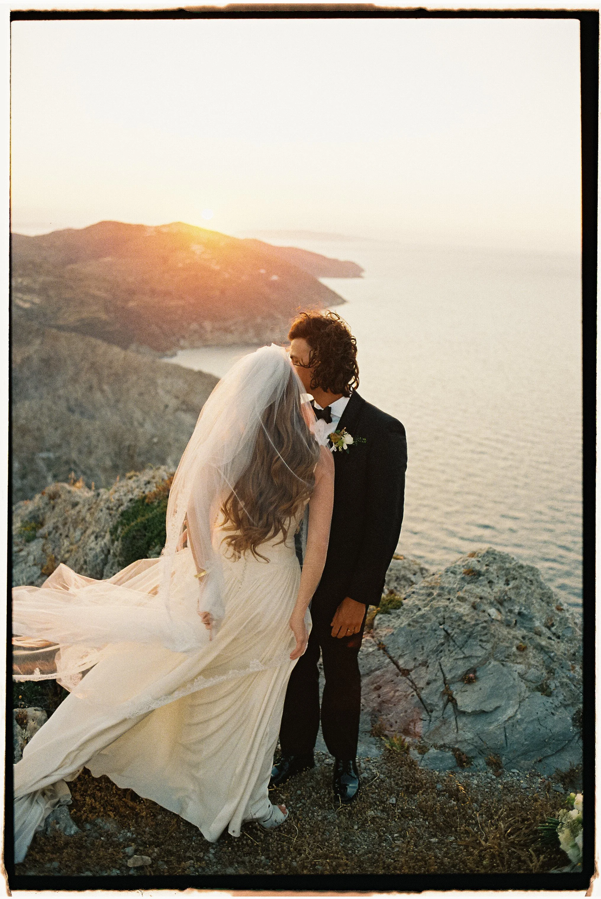 A bride and groom embracing on a rocky coastline at sunset, with the bride wearing a flowing white gown and veil, and the groom in a black tuxedo.