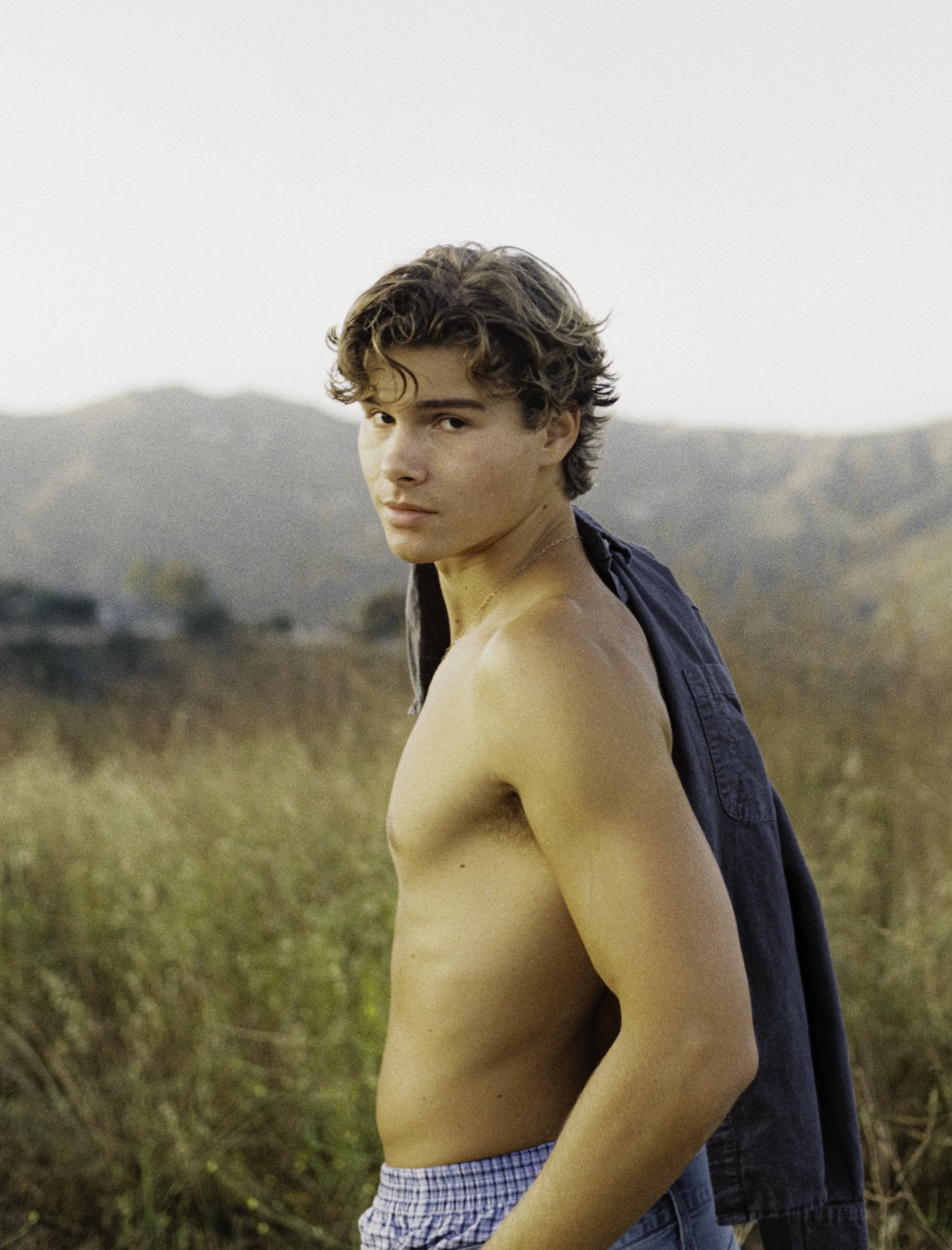 A young man with curly brown hair and a serious expression, shirtless with a blue shirt draped over his shoulders, standing outdoors in a grassy field with mountains in the background.