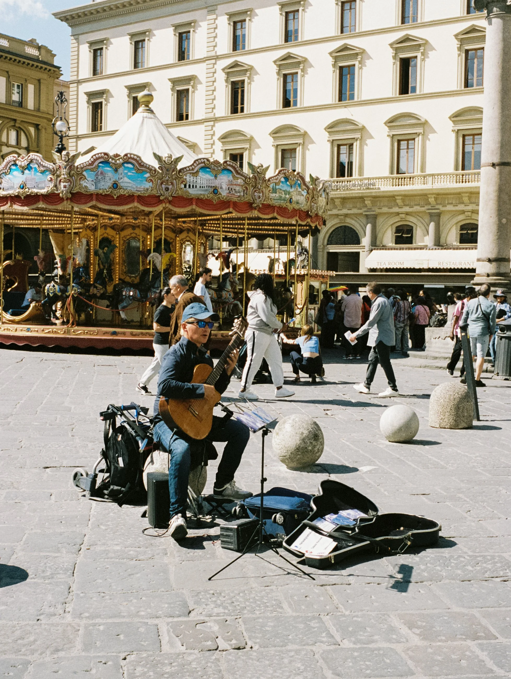 A street performer playing a guitar on a city square with a carousel and crowded pedestrians in the background.