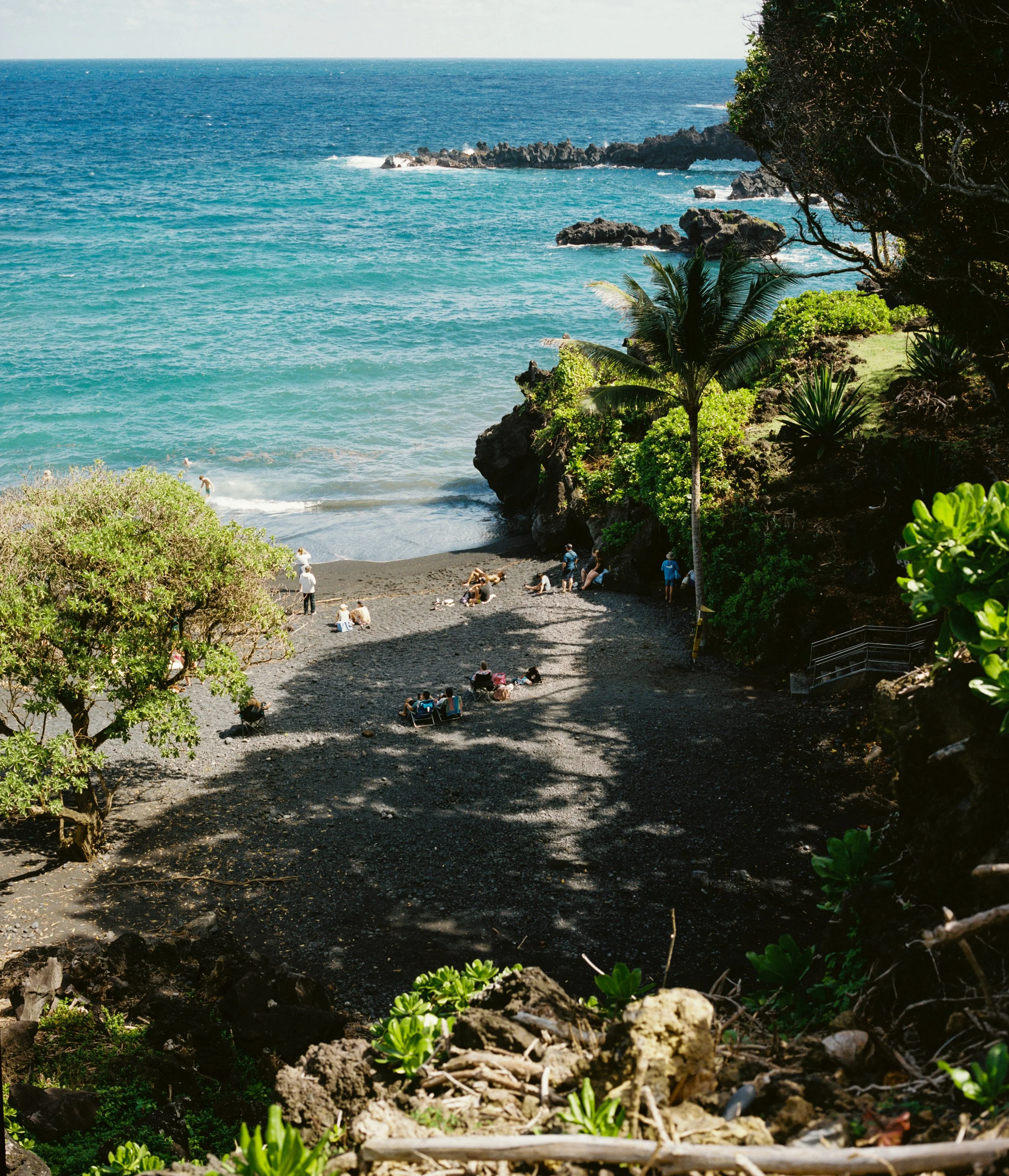 A beach with black sand and turquoise water, surrounded by tropical green plants and trees, with people relaxing and enjoying the scenery.