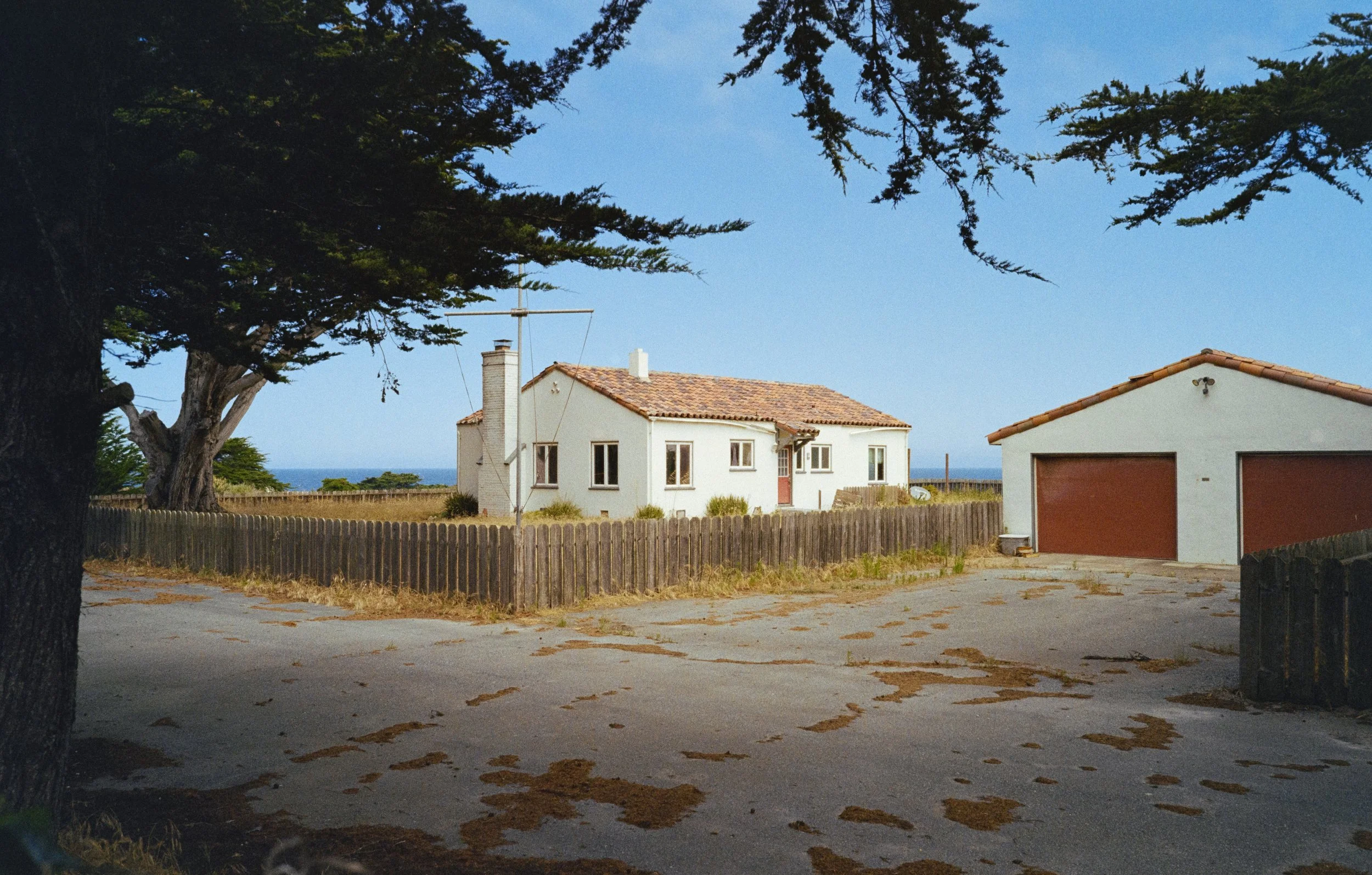 A white house with a tiled roof and multiple windows, surrounded by a wooden fence, with a detached garage featuring a red door, set outdoors with trees and a view of the ocean in the background.