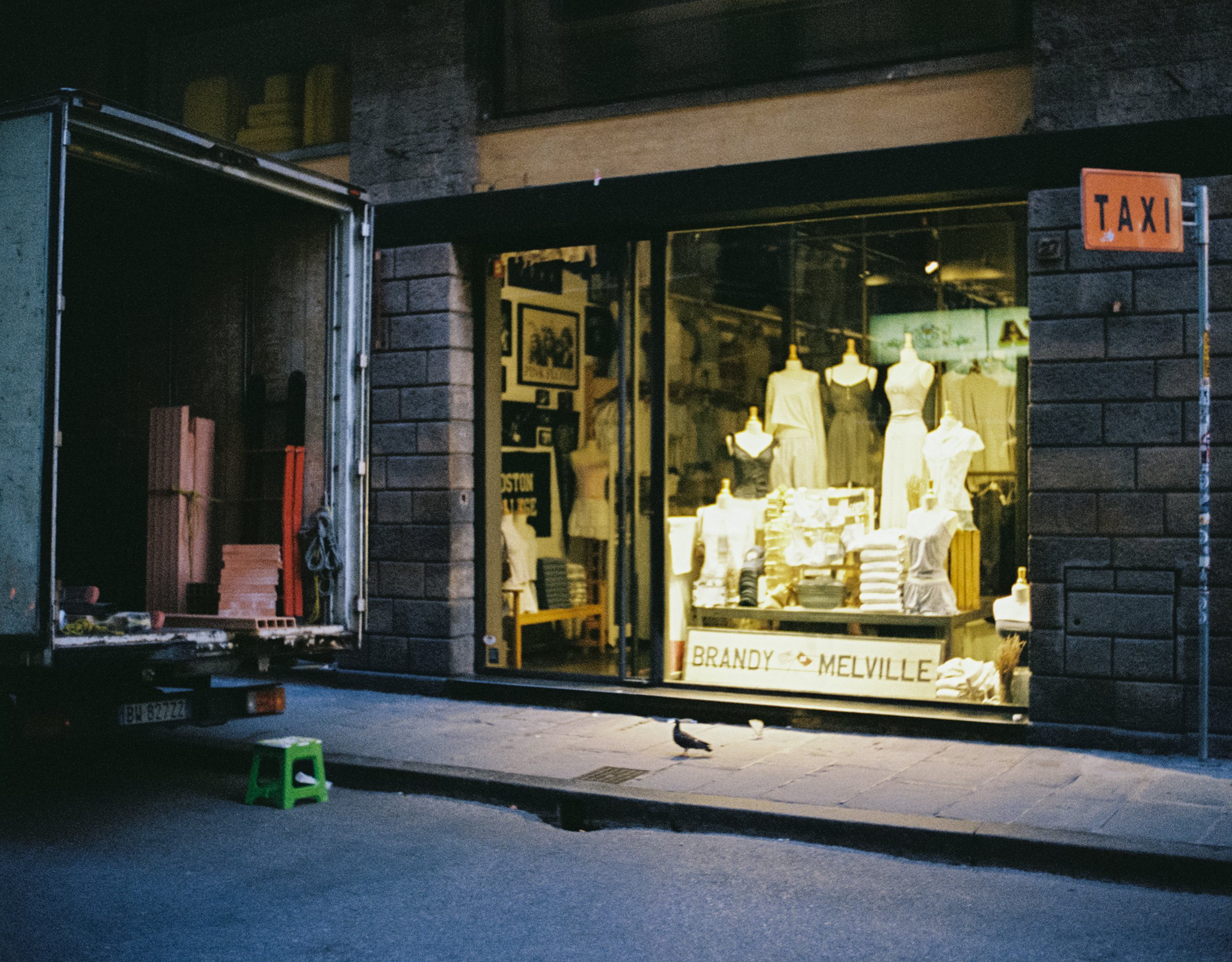 A clothing store with mannequins dressed in white and gray garments displayed in the window, and a sign reading 'BRANDY MELVILLE', with a delivery truck parked nearby and a few pigeons on the sidewalk.