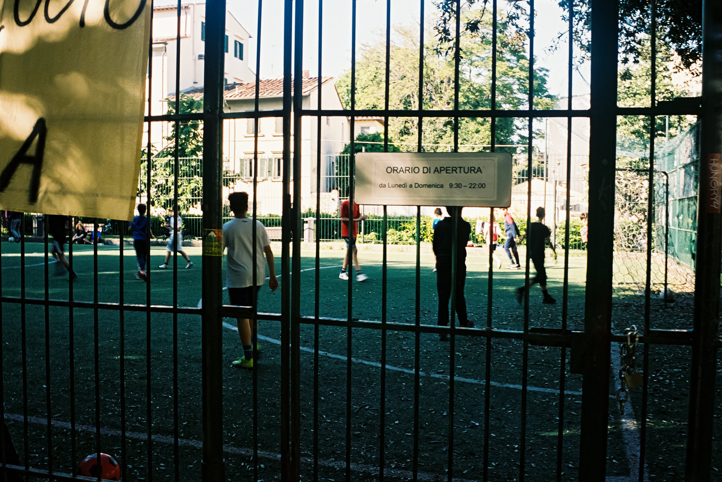 Children and teenagers playing soccer on a fenced outdoor sports court during daytime, with buildings and trees in the background.