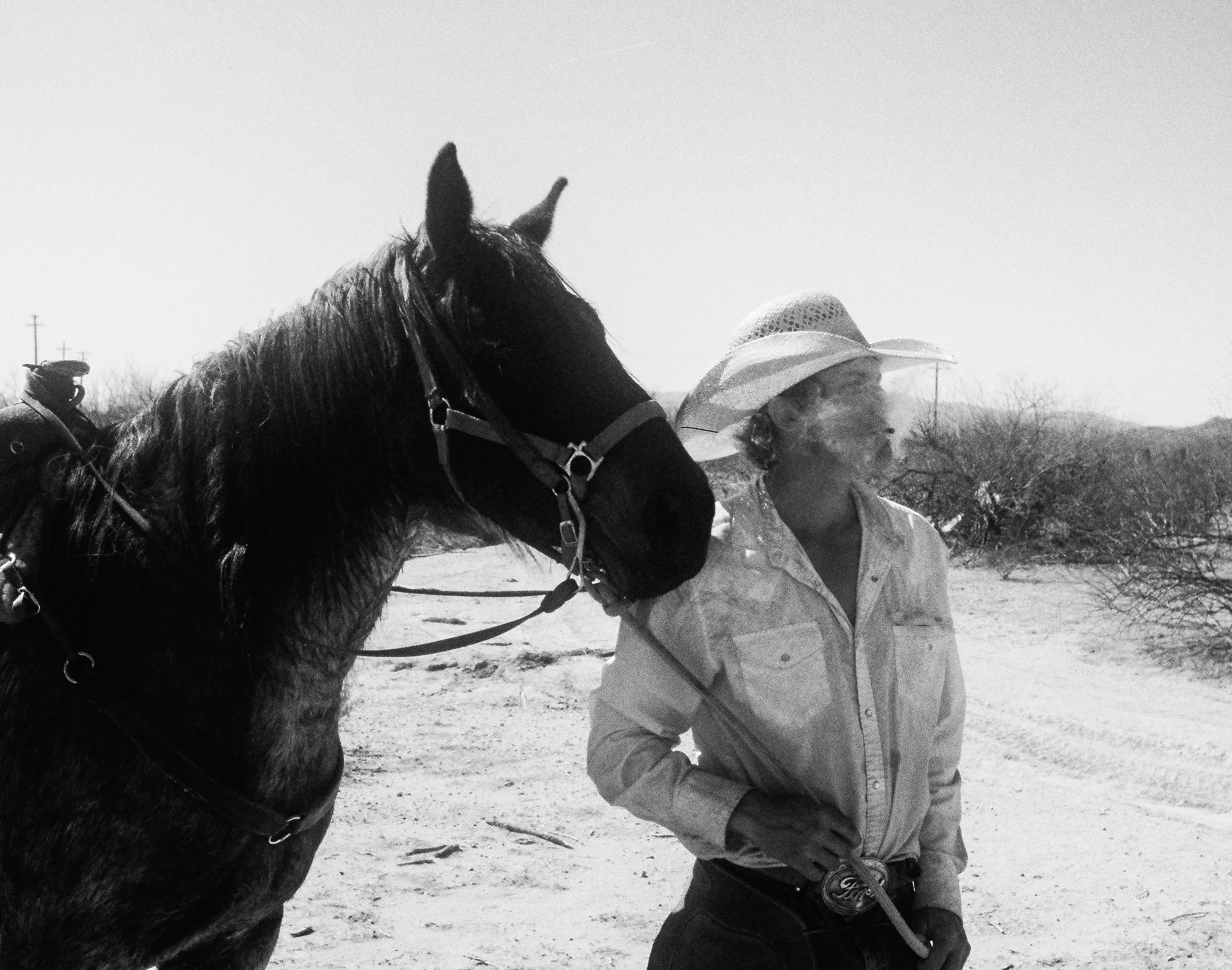 A person wearing a wide-brimmed hat and jacket standing next to a horse in a desert landscape with sparse vegetation.