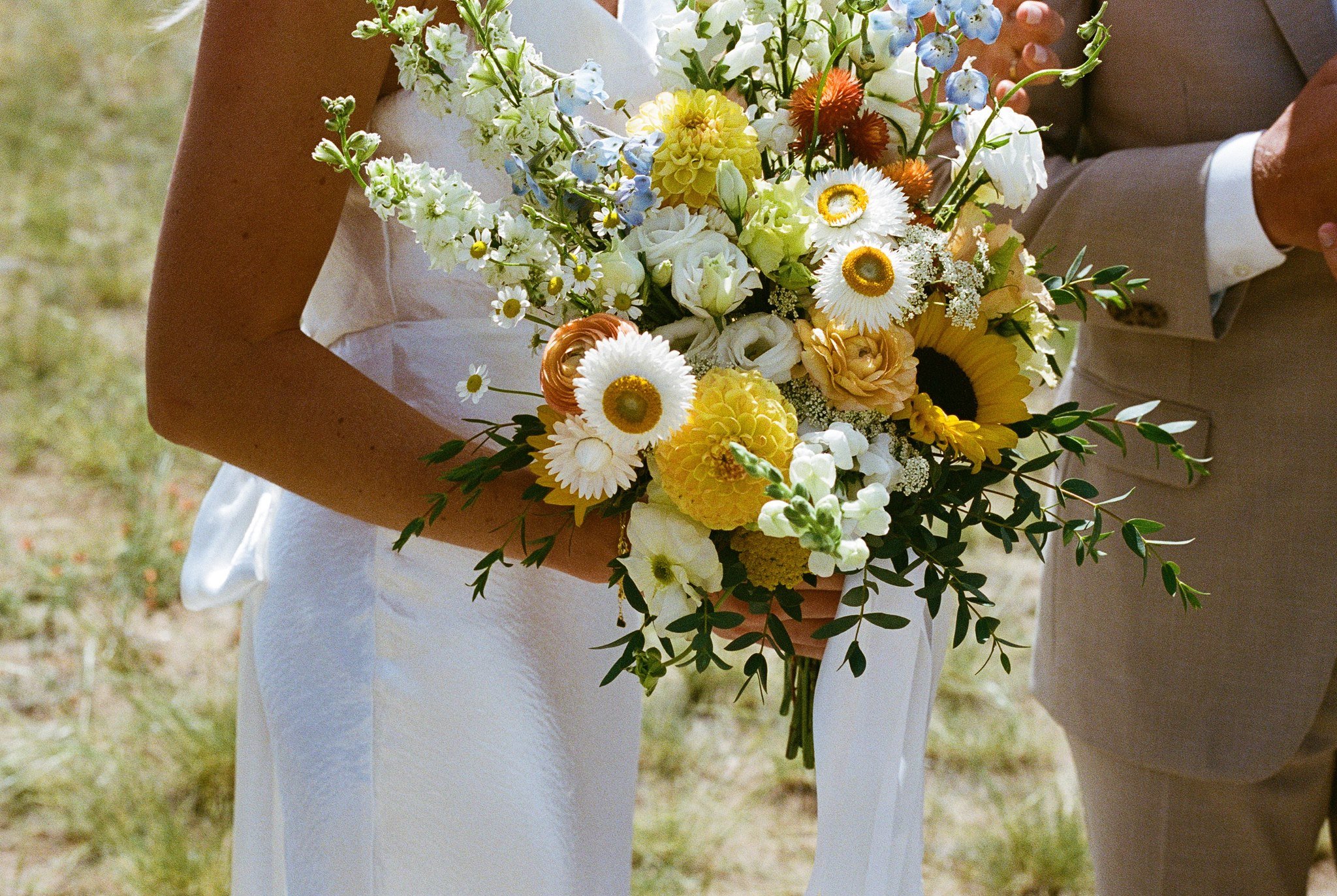 A bride and groom holding a bouquet of mixed flowers including daisies, sunflowers, and other colorful blooms outdoors.