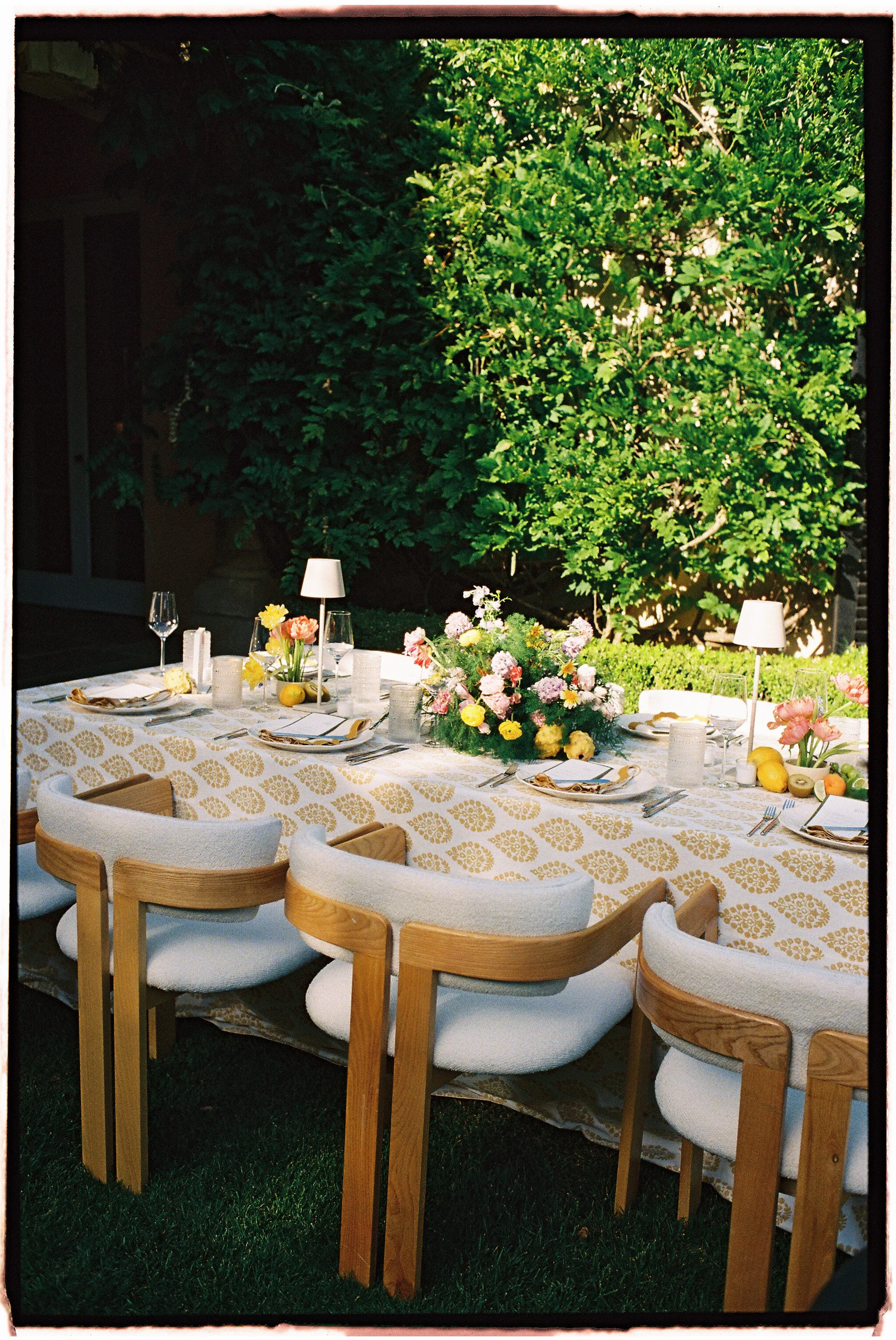 Outdoor dining table decorated with flowers, candles, and tableware, set on a grassy area with lush green bushes in the background.