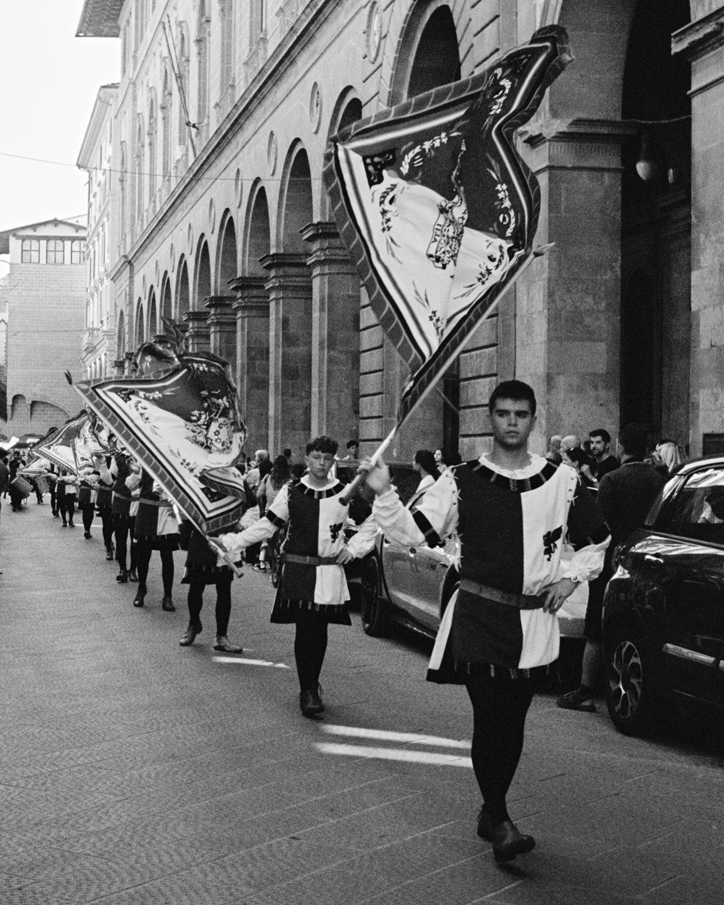 Group of people dressed in historical costumes, carrying flags in a parade on a city street.