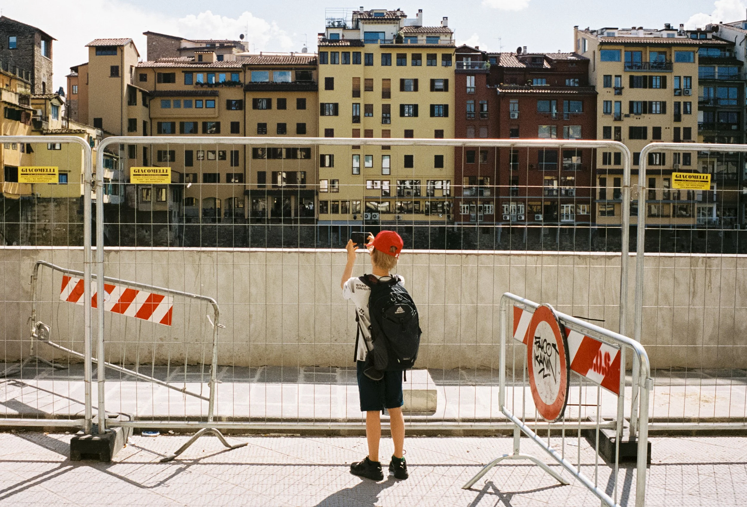 A boy with a red cap and a backpack is taking a photo with a smartphone in front of a construction site with metal barricades, graffiti, and apartment buildings in the background.
