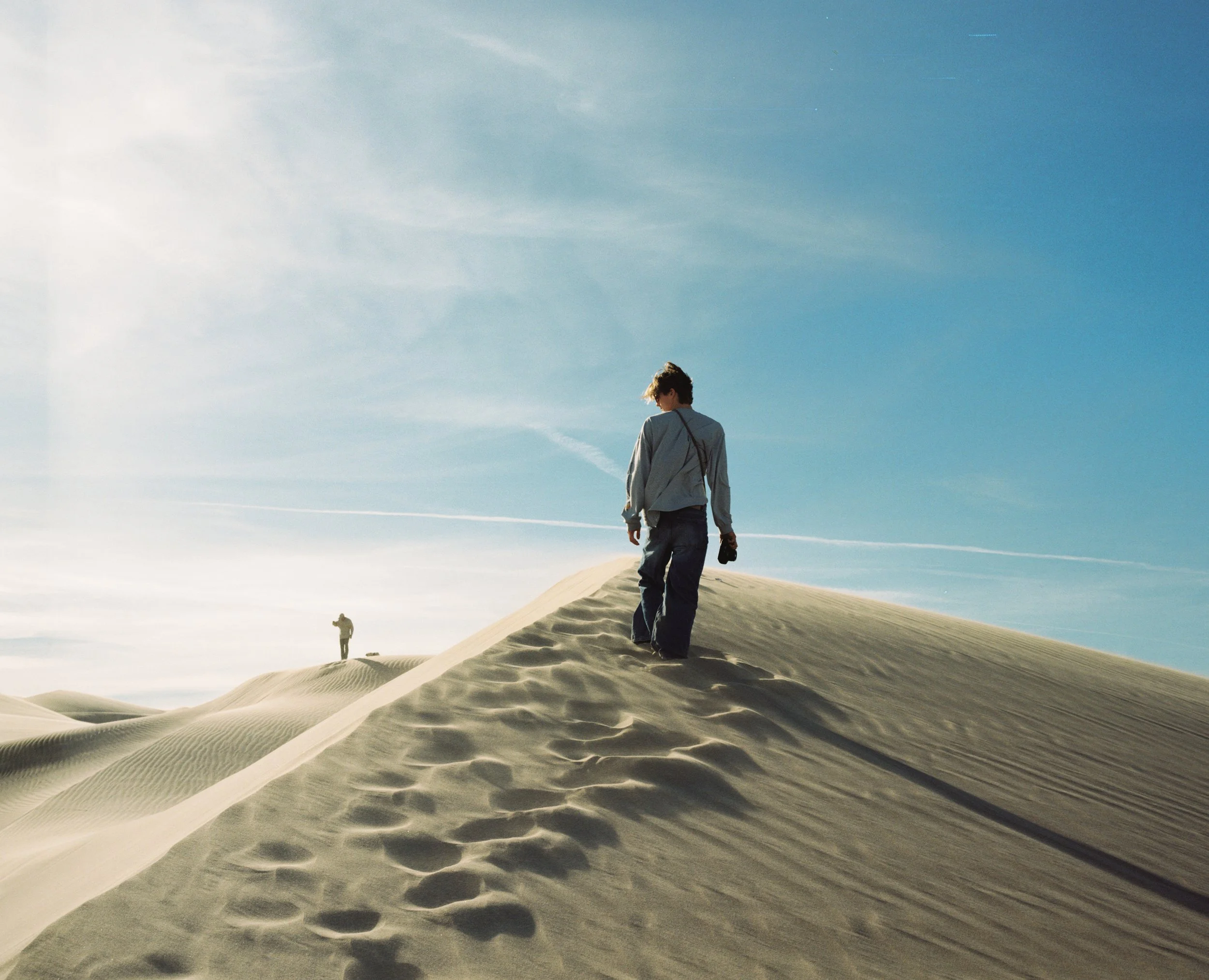 A person walking on sand dunes with footprints visible, under a clear blue sky with some clouds.