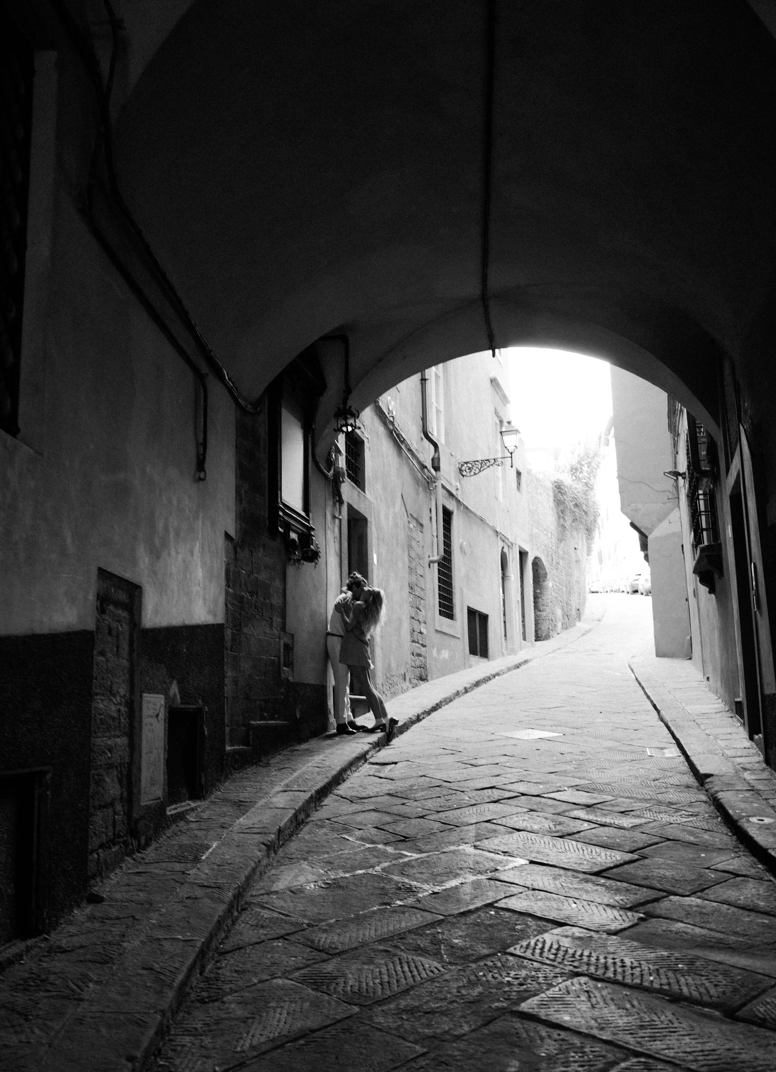 A black and white photograph of a narrow European street with cobblestone paving, an archway overhead, and a romantic couple embracing in the distance.