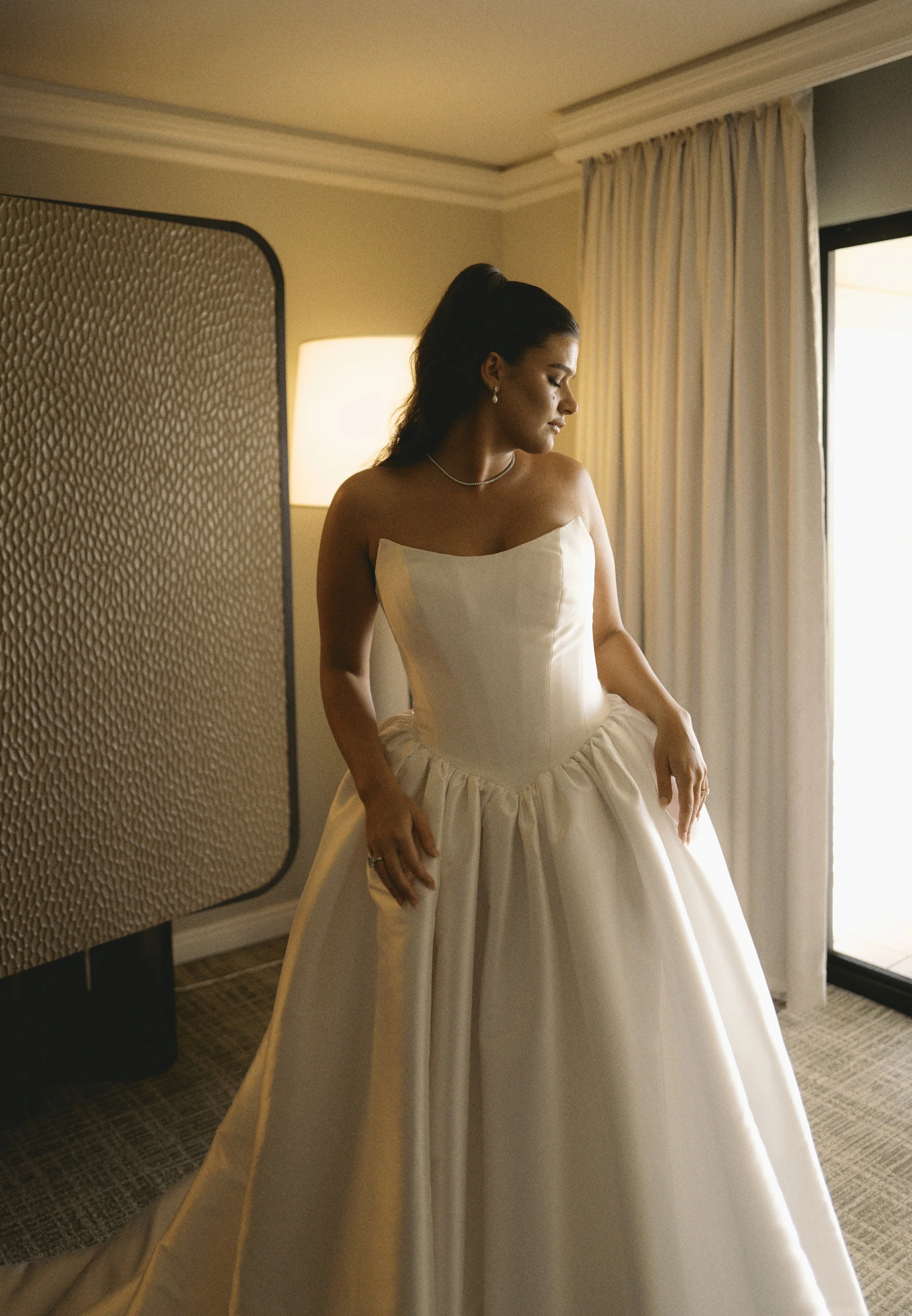 A woman dressed in a strapless white wedding gown standing near a window with curtains, looking down with a contemplative expression in a warmly lit room.