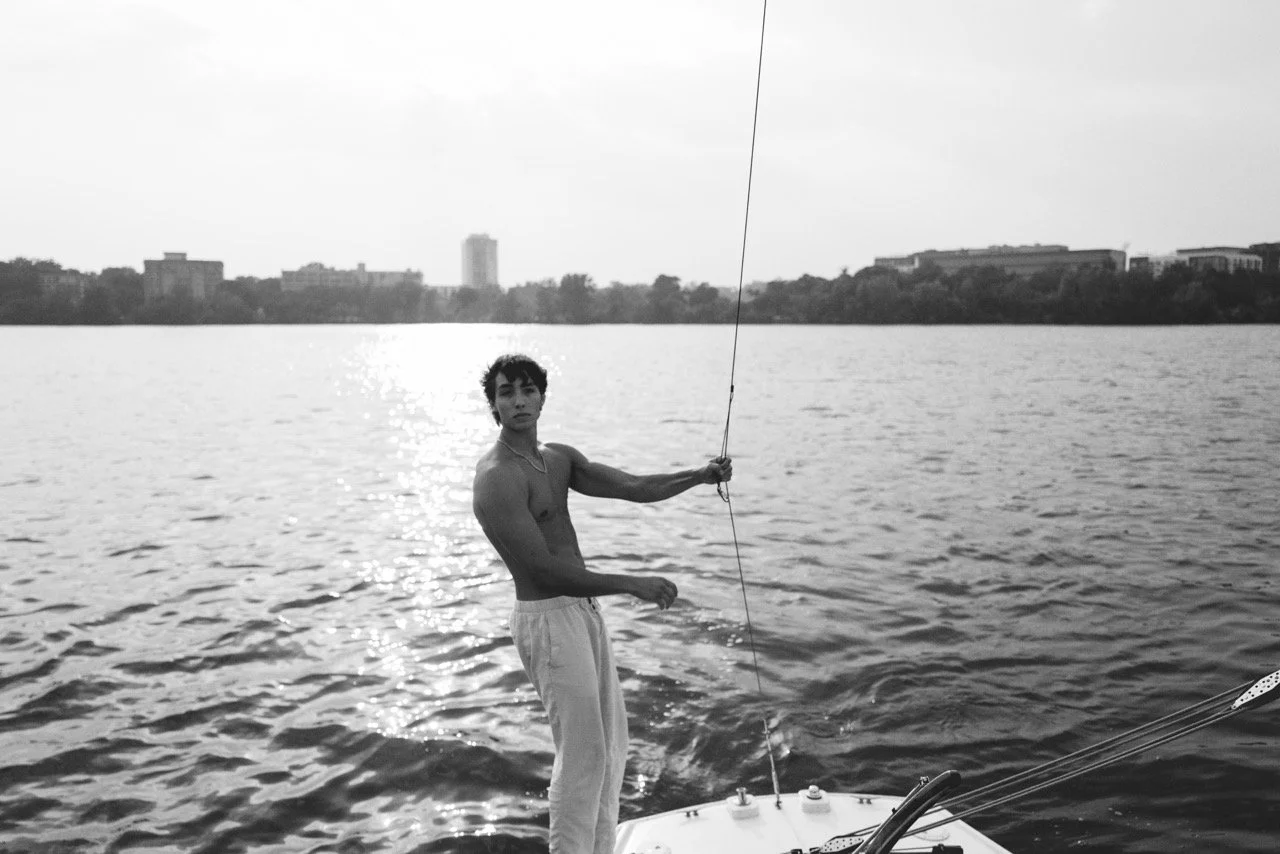 A shirtless young man standing on a boat while holding a fishing rod in a large body of water with city buildings in the background.