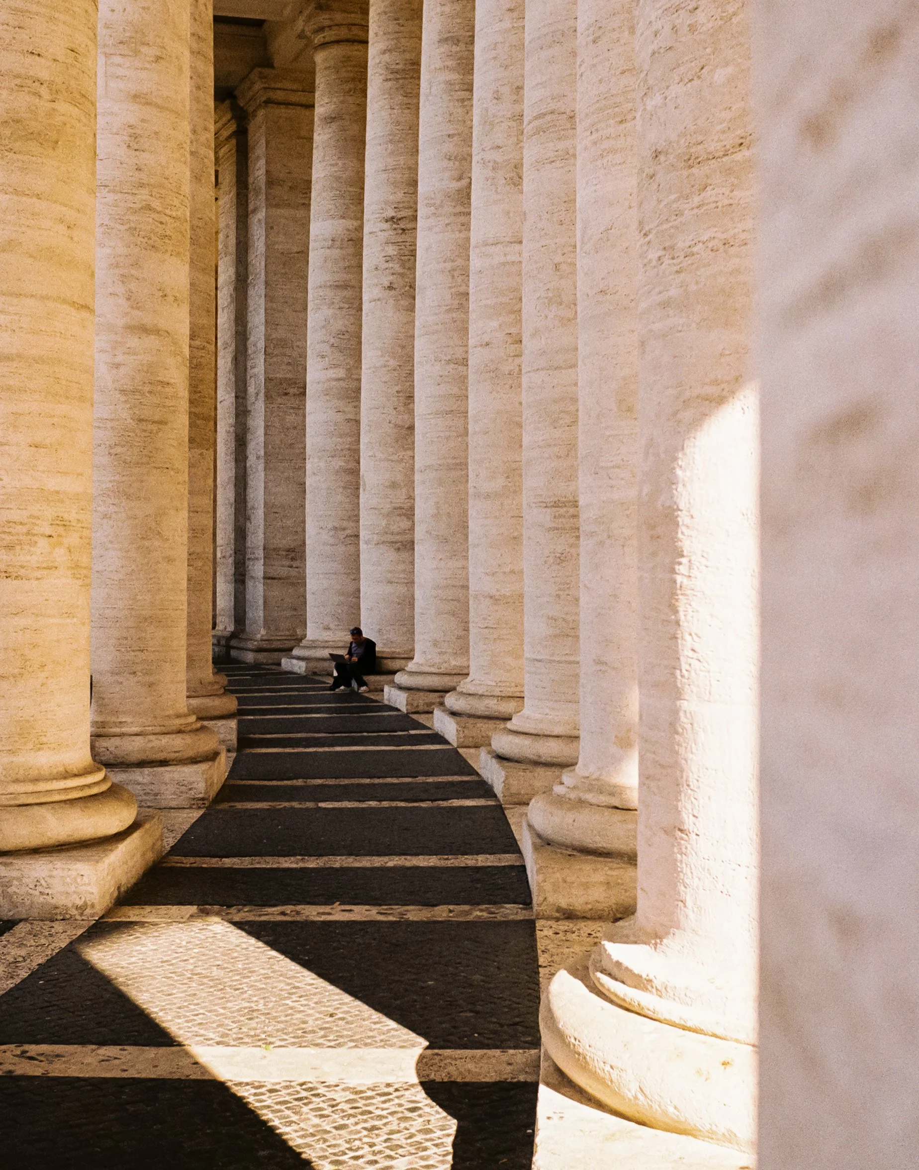 A person sitting on a bench between large white stone columns in a classical architectural setting, with sunlight creating shadows on the ground.