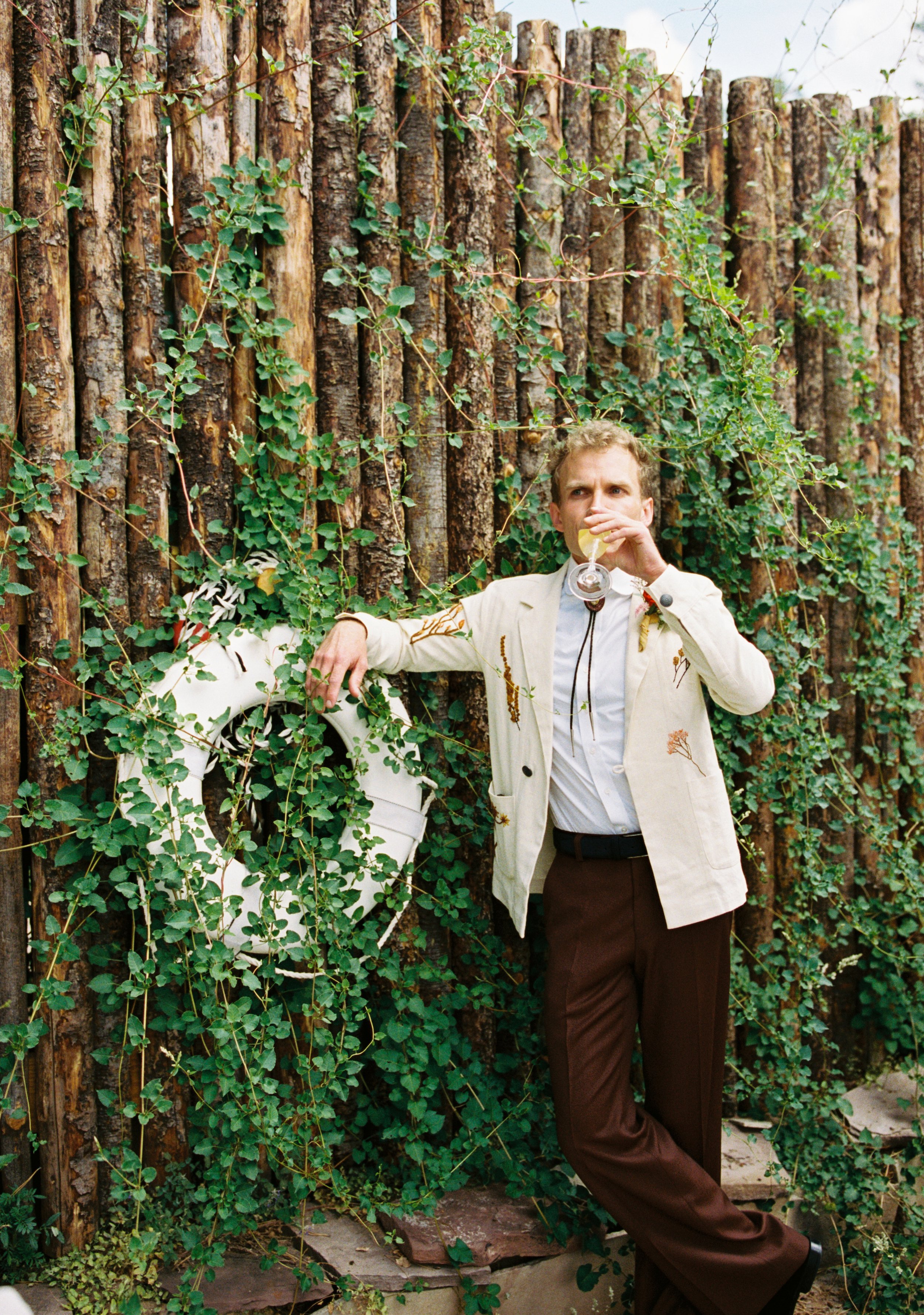 A man in a beige jacket and brown pants stands against a wooden fence covered with green ivy, sipping a drink with a lemon slice.