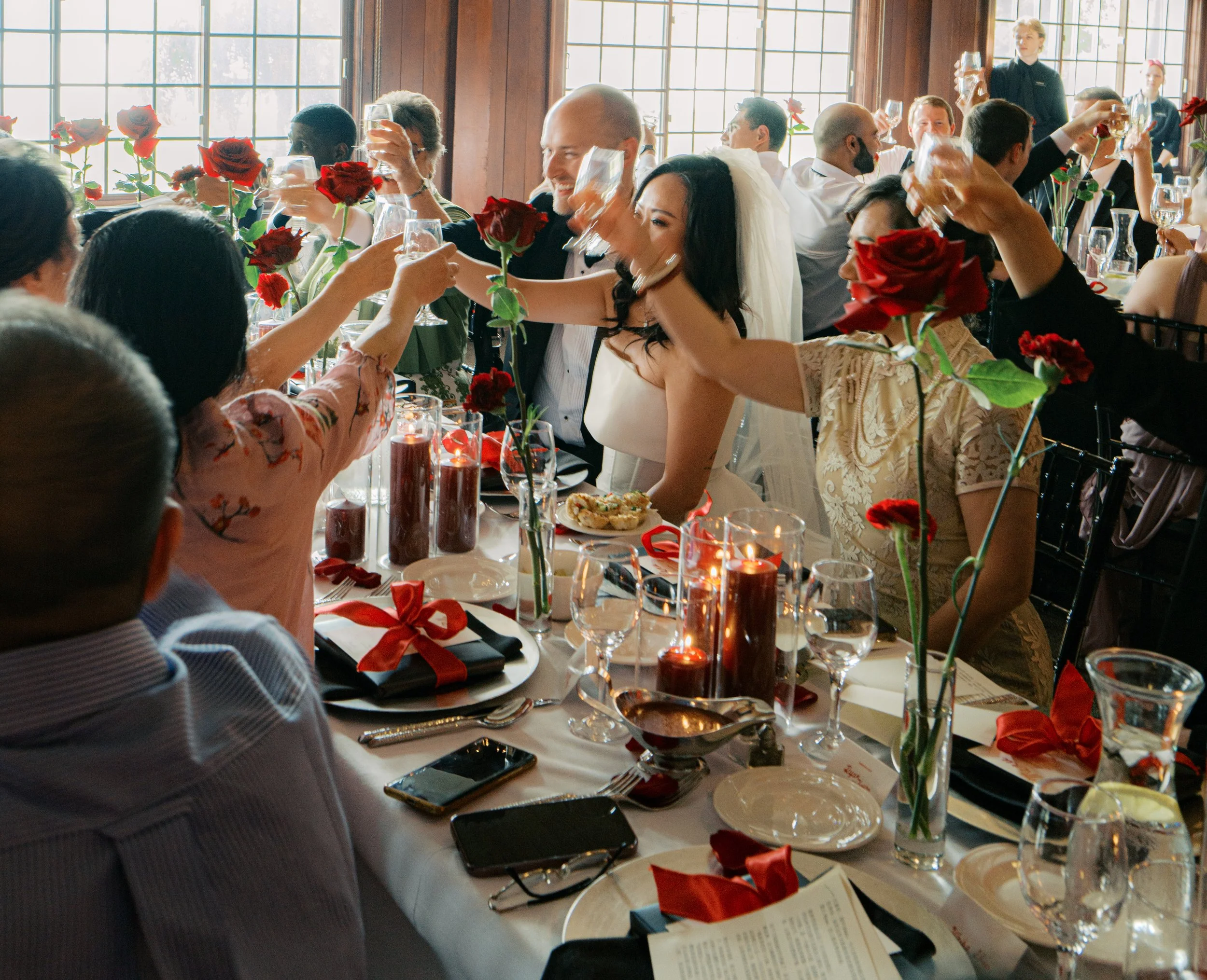 People celebrating at a wedding reception with roses and glasses of wine, seated at a long table with candles, plates, and cutlery.