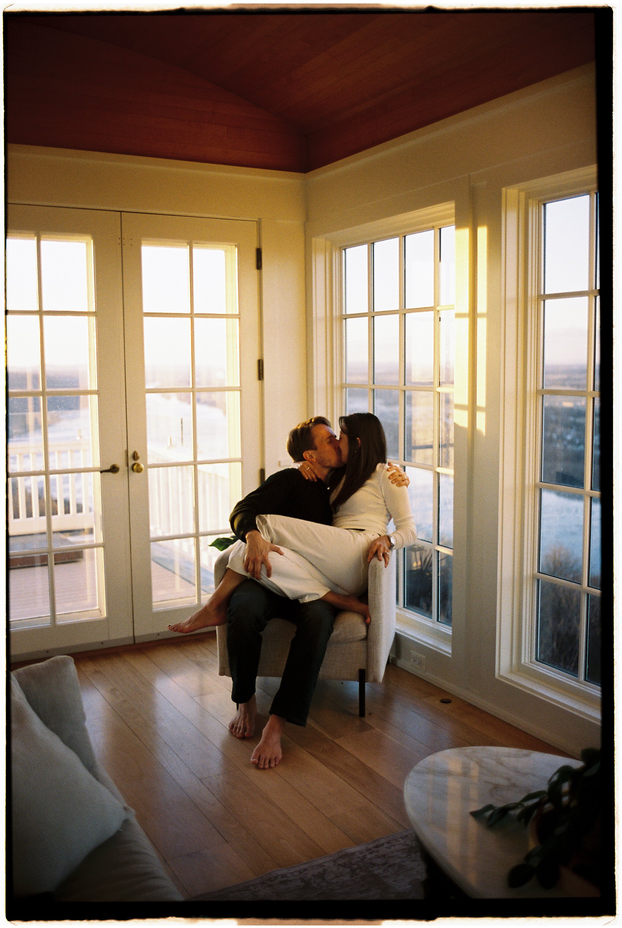 A couple sharing a kiss in a bright living room with large windows and wooden floors, sunlight streaming in.