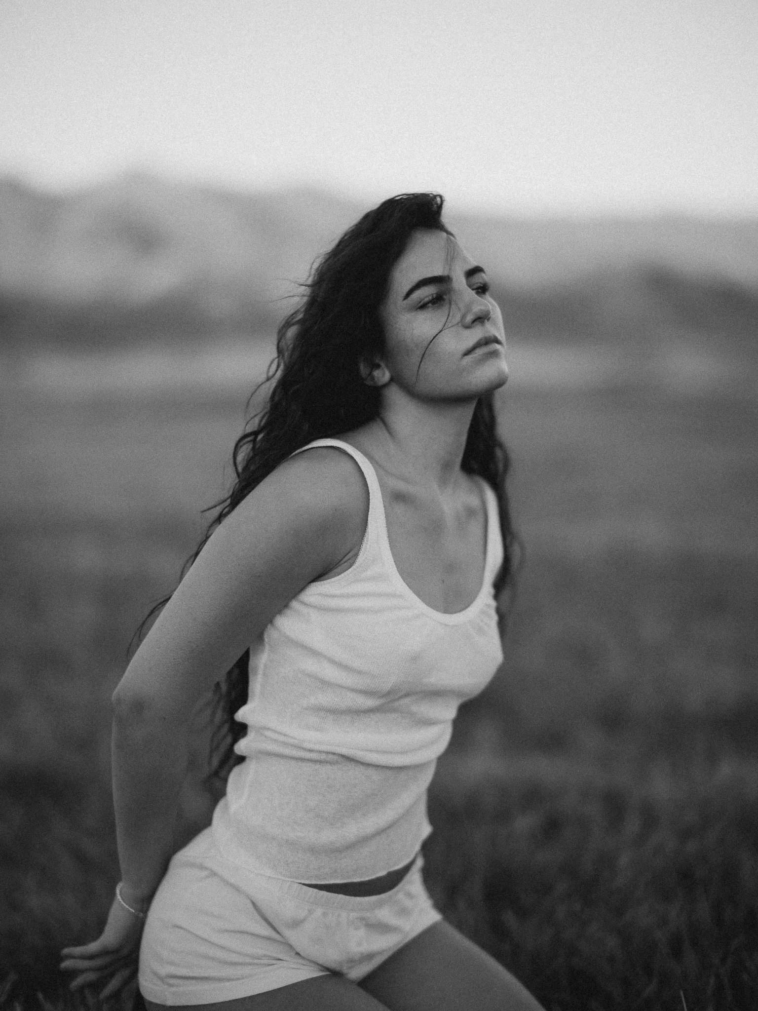 A young woman in a white tank top and shorts sitting outdoors on grass, with her back slightly arched, gazing thoughtfully into the distance. The photo is in black and white.