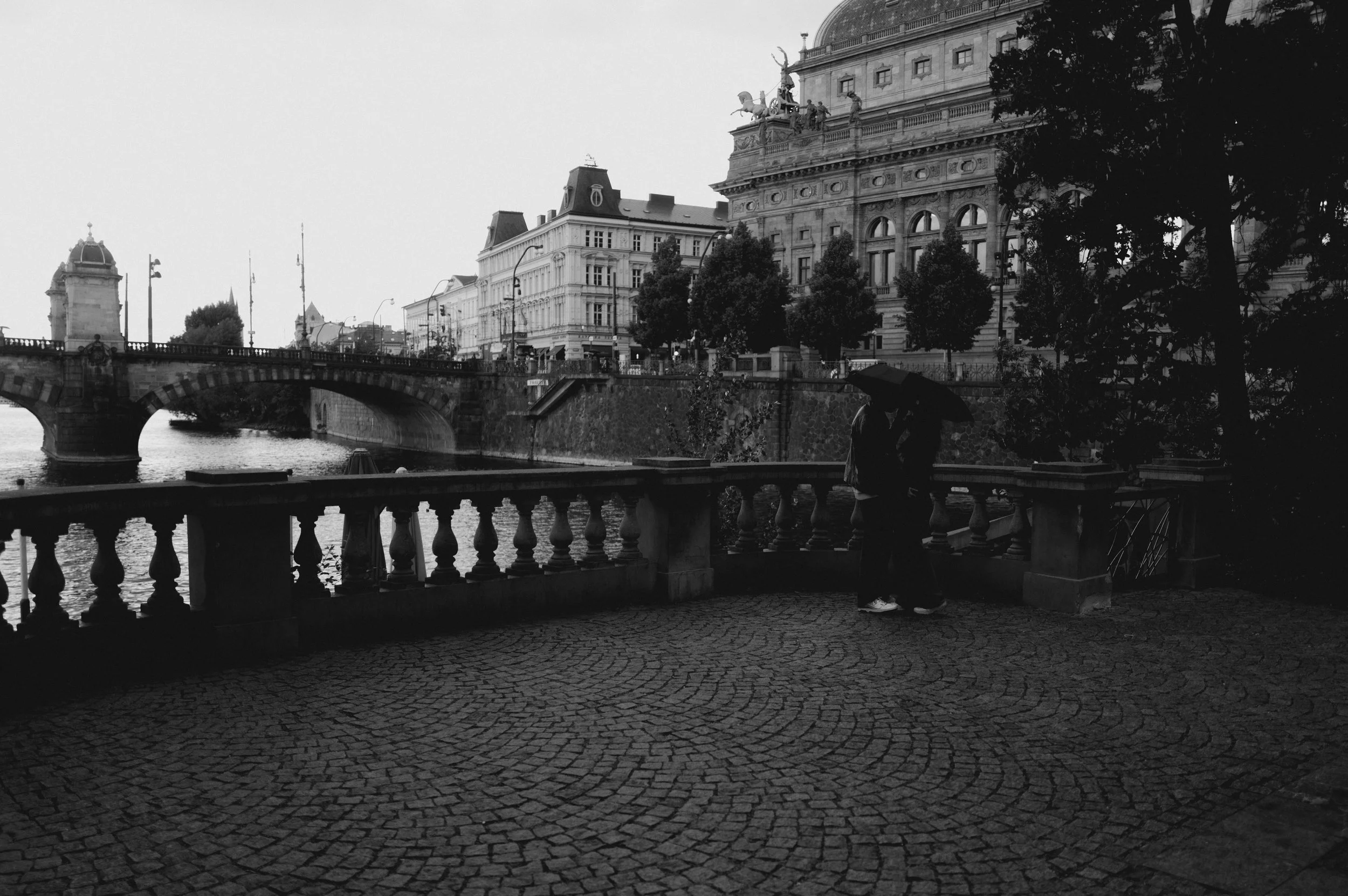A couple standing close with umbrellas overlooking a river, with historic buildings and a bridge in the background, on a rainy day.