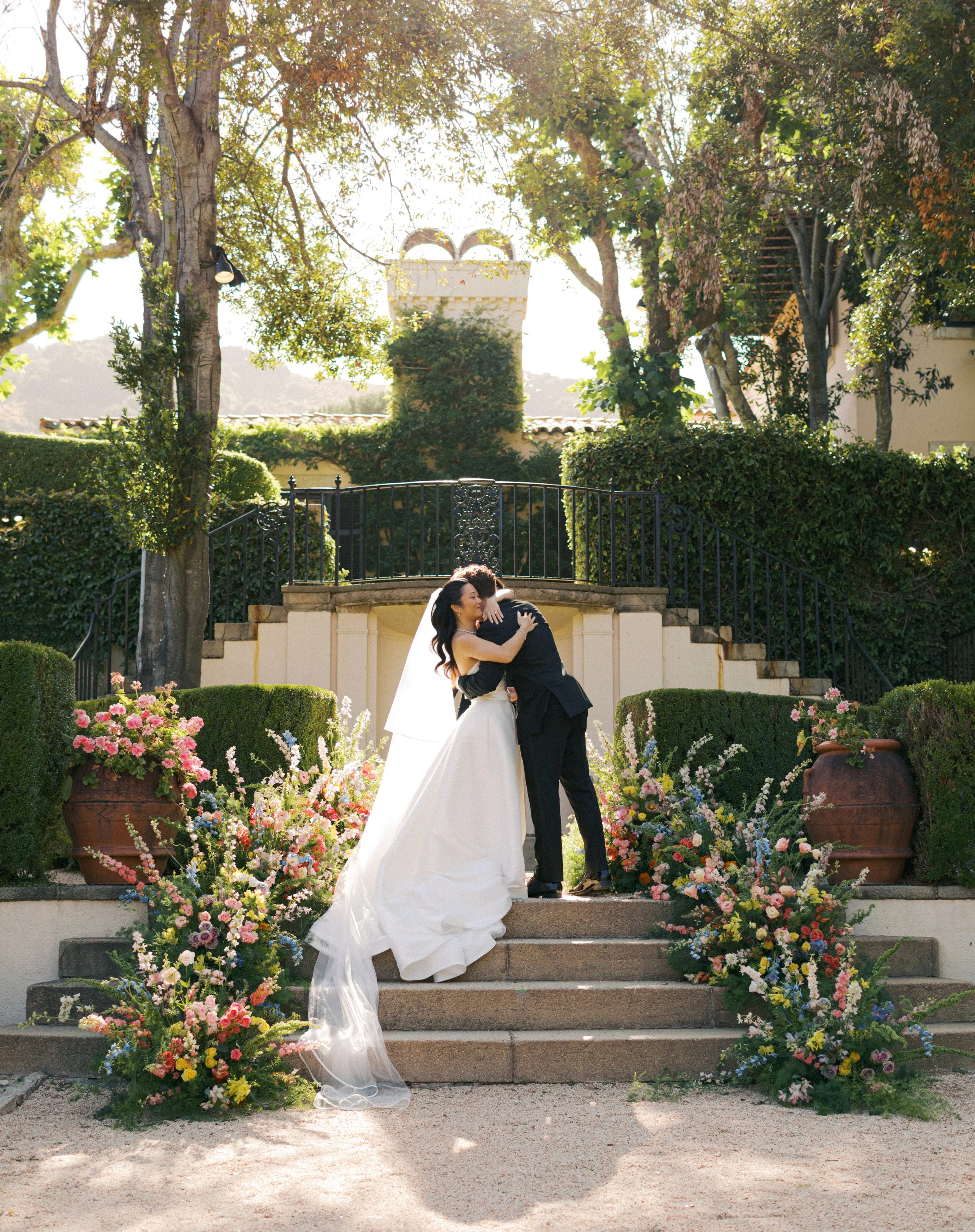 A bride and groom sharing an embrace on outdoor wedding stairs. The bride wears a white wedding gown and veil, and the groom is dressed in a dark suit. The stairway is decorated with colorful flowers in large pots and surrounded by lush bushes and ta