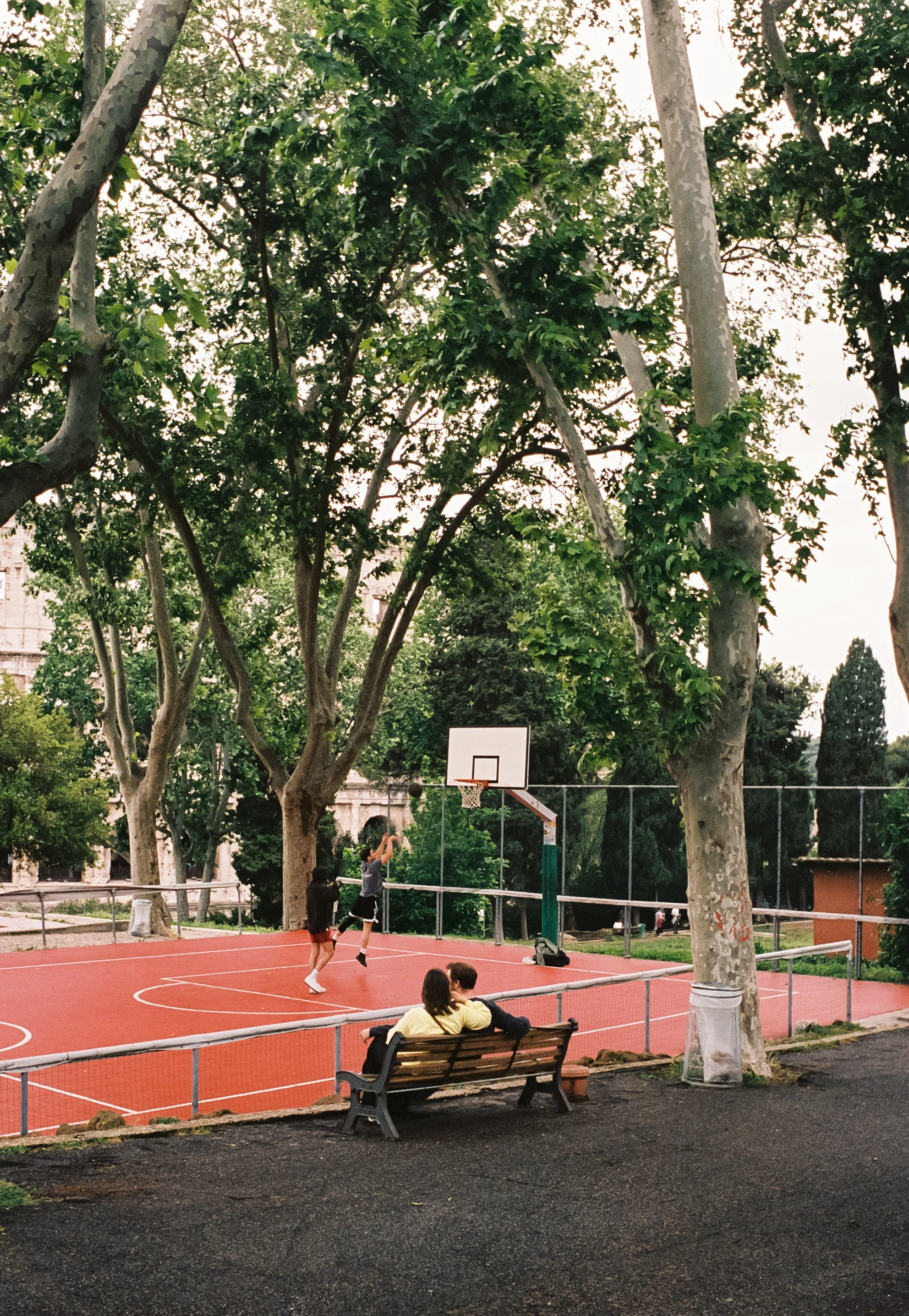 Two children playing basketball on an outdoor court, while a person sits on a park bench watching, surrounded by tall trees.