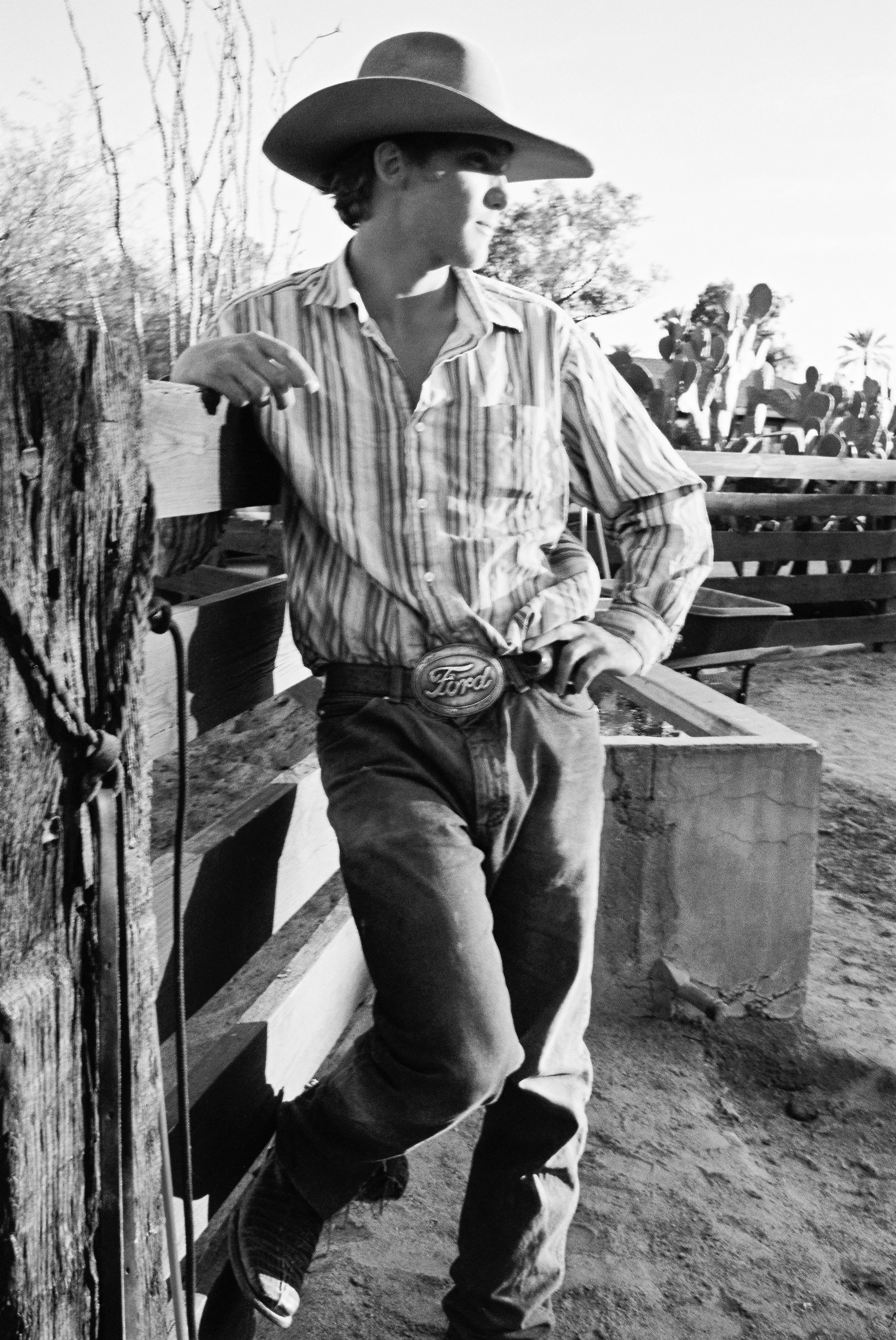 A young man dressed as a cowboy, wearing a wide-brimmed hat, striped shirt, jeans, and a large belt buckle, leaning against a wooden fence in a rural setting with trees and cacti in the background.