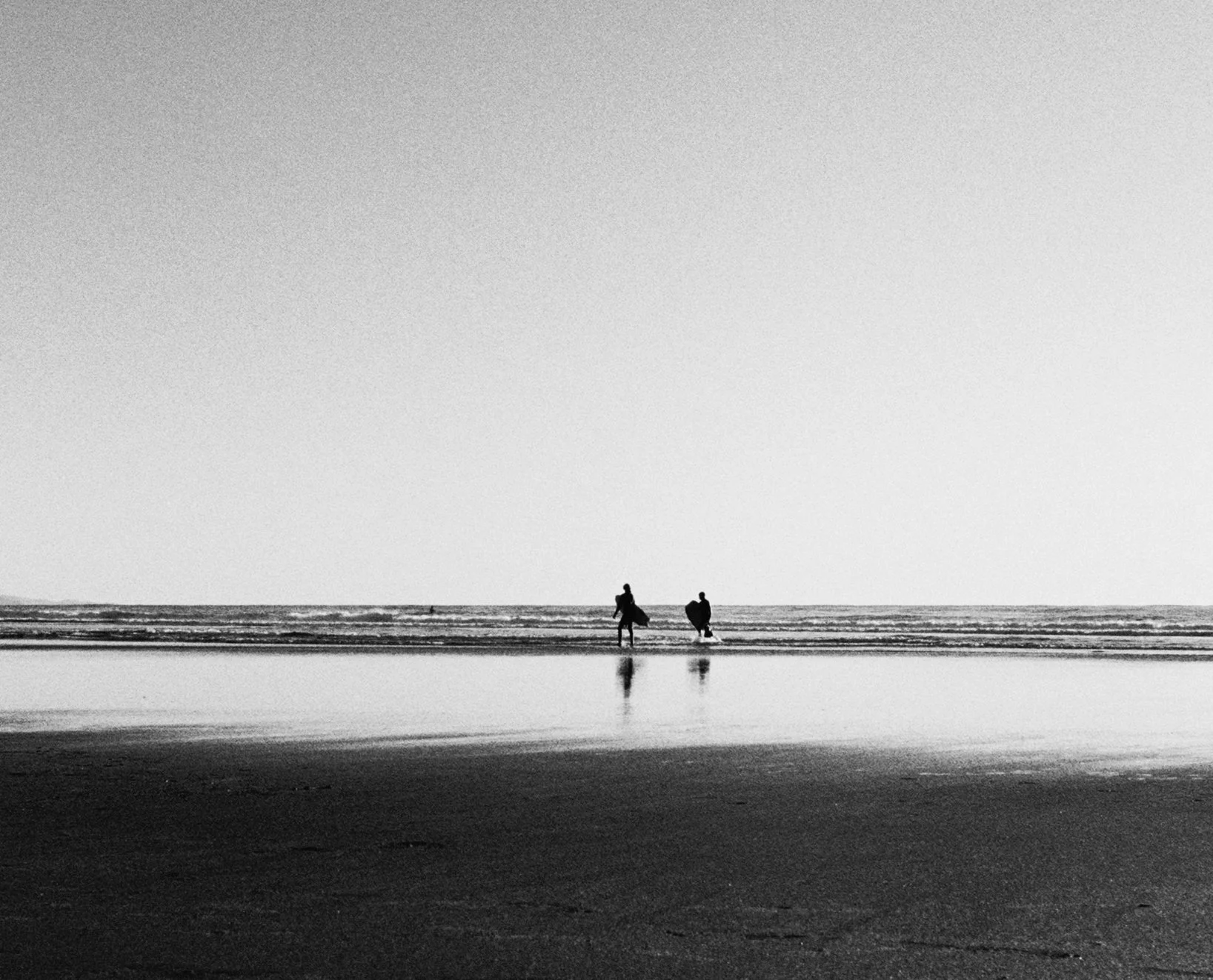 Black and white photo of two people with surfboards walking into the ocean on a beach, with reflections on wet sand in the foreground.