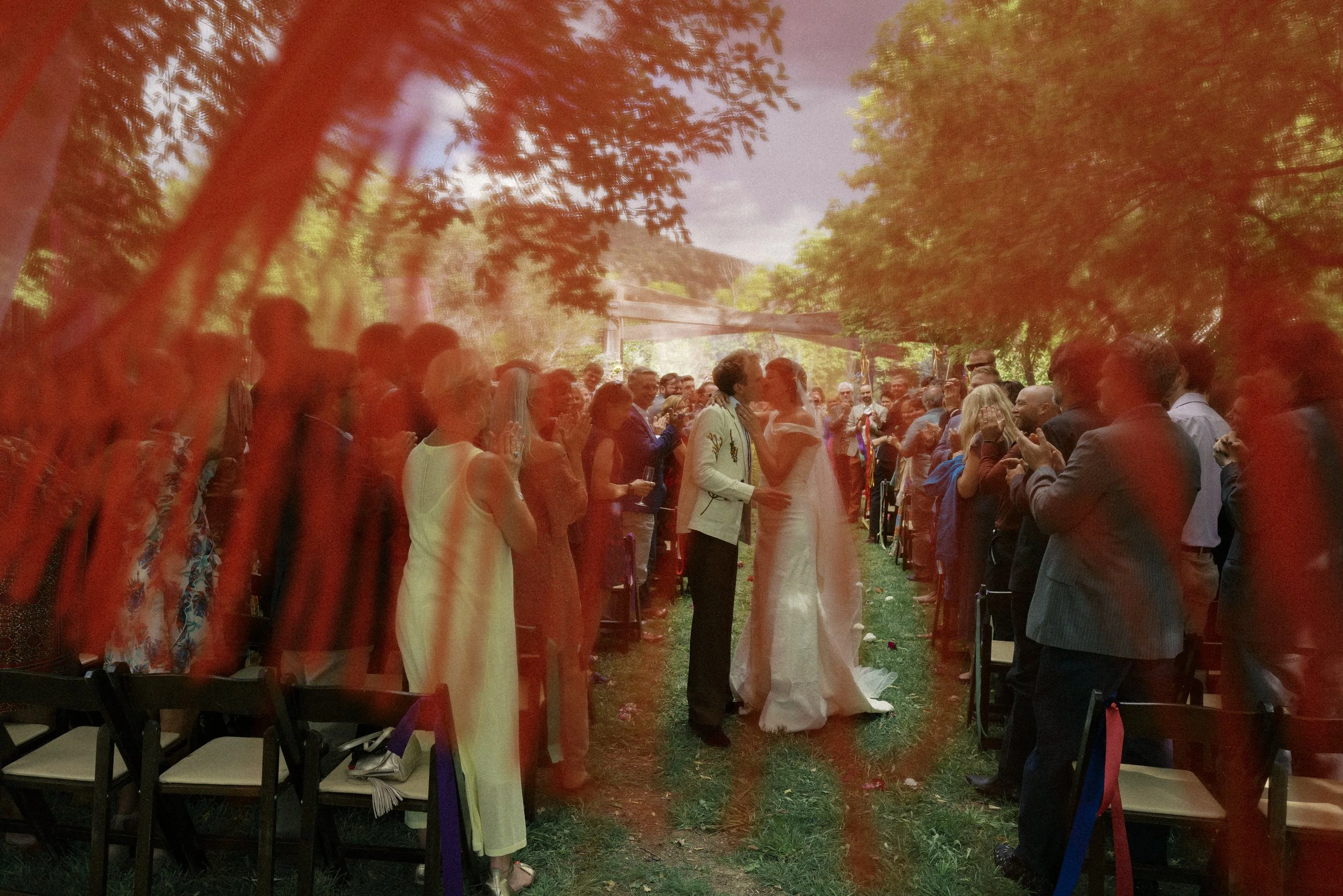 A wedding ceremony taking place outdoors with a bride and groom at the center, surrounded by guests standing and facing each other, under trees with sunlight filtering through.