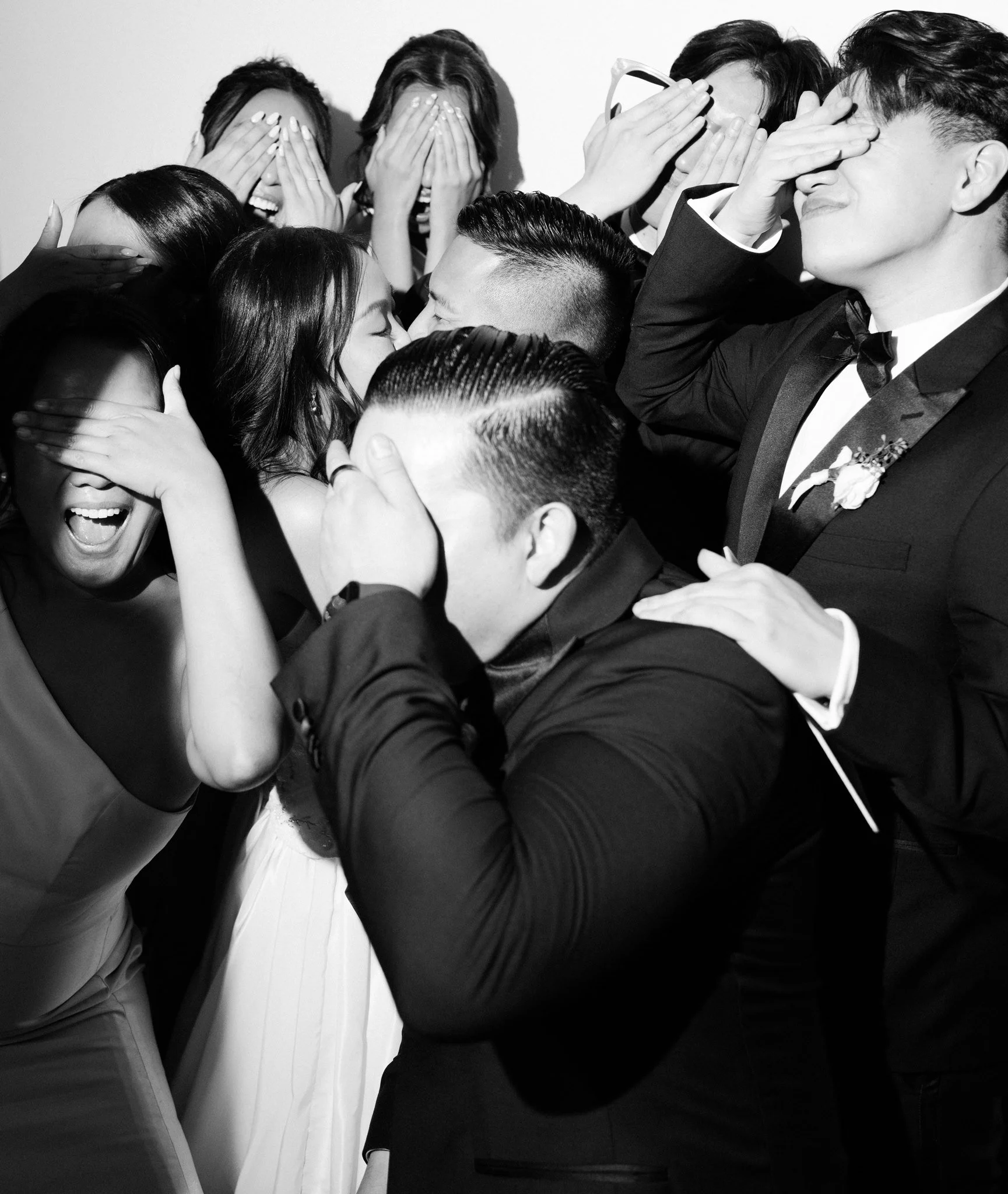 A group of people dressed in formal attire, covering their eyes with their hands and leaning in for a kiss, at a celebration or event.