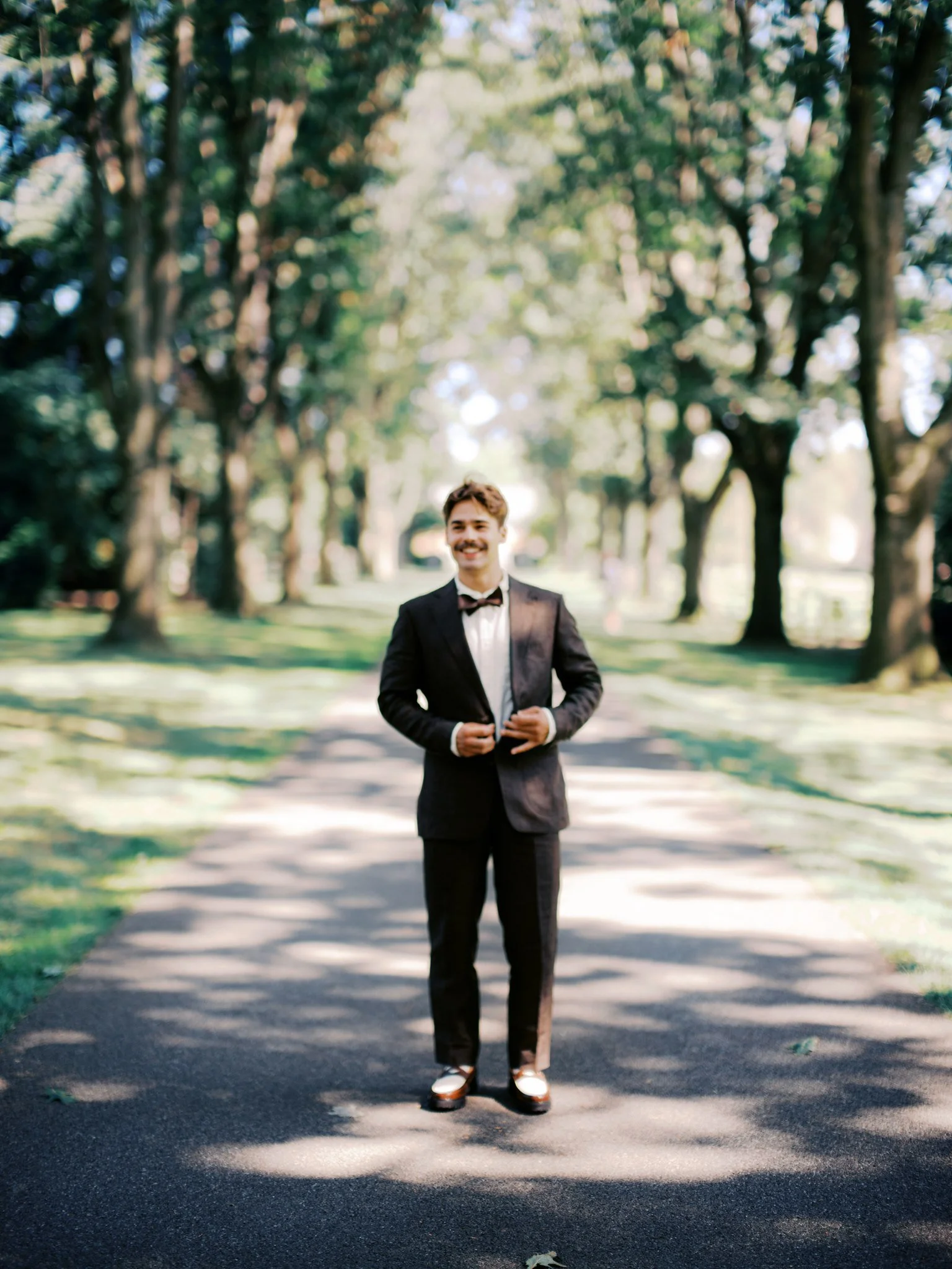 A man in a black tuxedo with a bow tie is standing on a paved path in a park with trees in the background. He is smiling and adjusting his jacket.