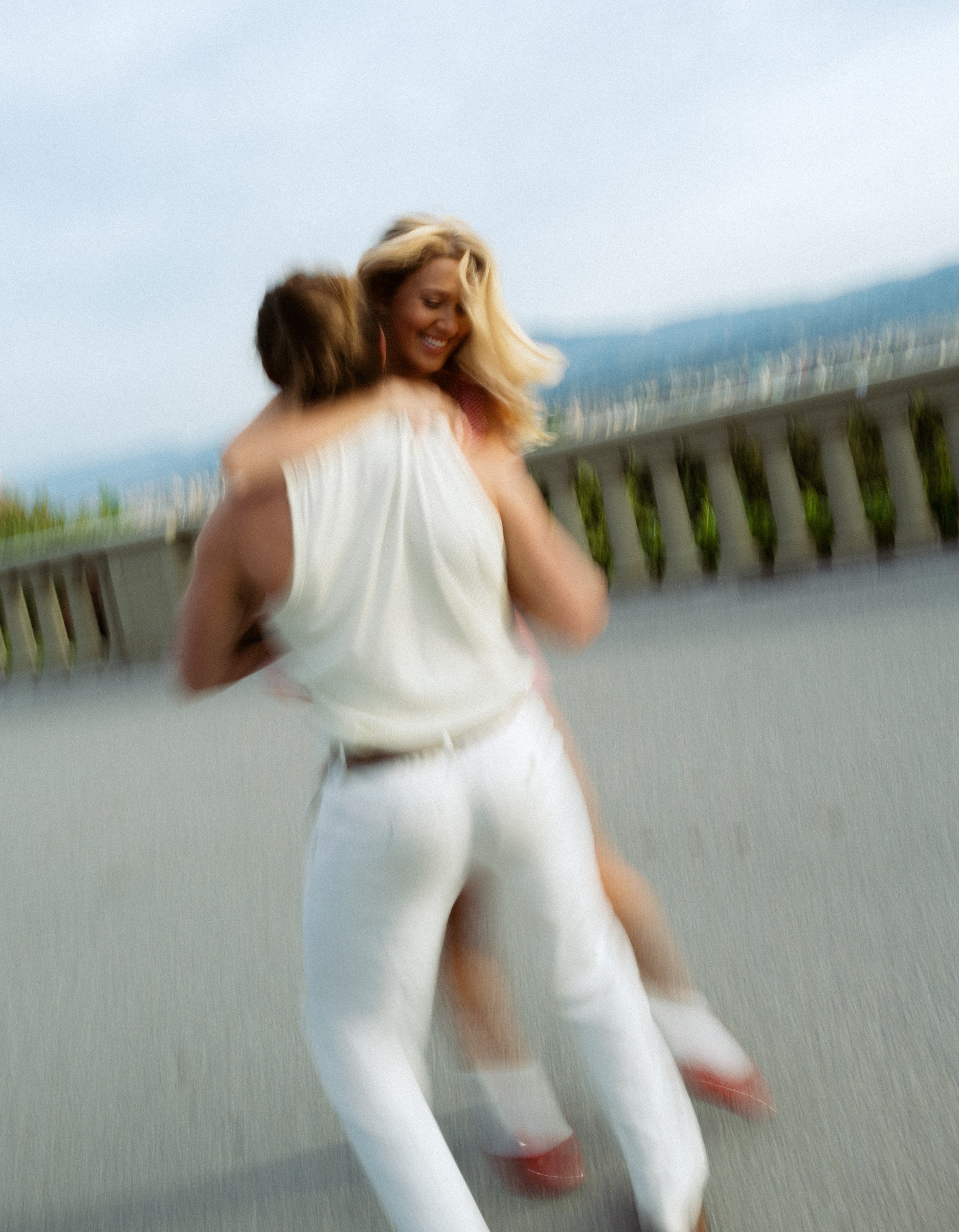 Two women embracing, one with blonde hair wearing a white top and the other with brown hair, outdoors on a balcony or terrace, with mountains and cityscape in the background, during daytime.