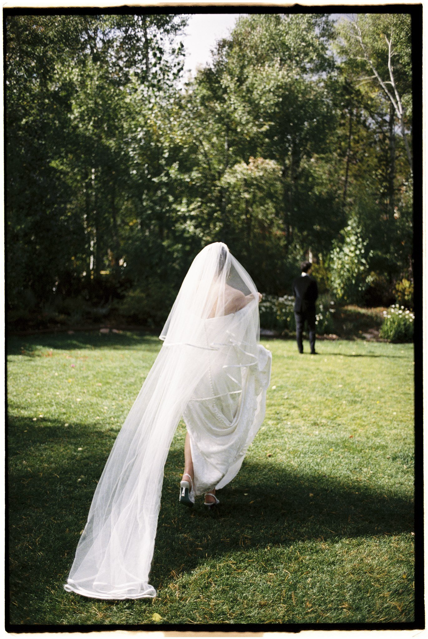 Bride walking on grass in wedding dress with veil, photo taken from behind, with a groom standing in the distance in an outdoor garden setting.
