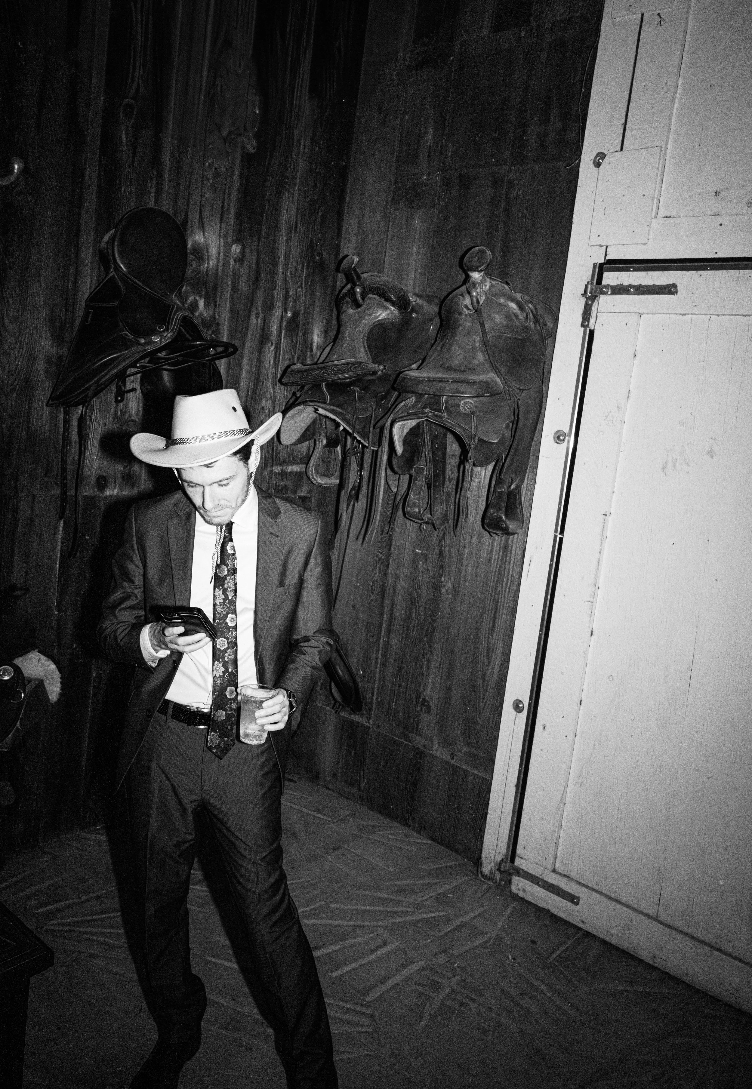 A man dressed in a suit with a floral tie and a cowboy hat is holding a drink and looking at his phone in a dimly lit room with wooden walls. There are two saddles hanging on the wall behind him.