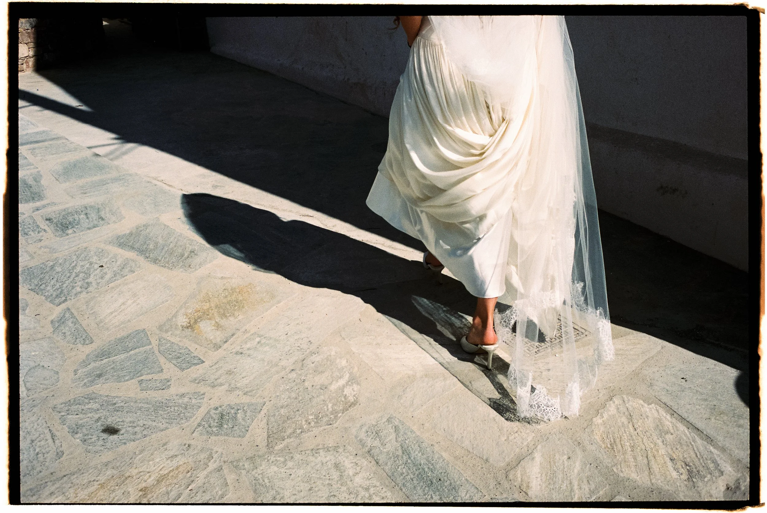 The image shows a woman in a flowing white dress with lace details at the bottom, standing on a patterned stone floor with her back to the camera. She appears to be stepping forward, with sunlight casting her shadow on the ground.