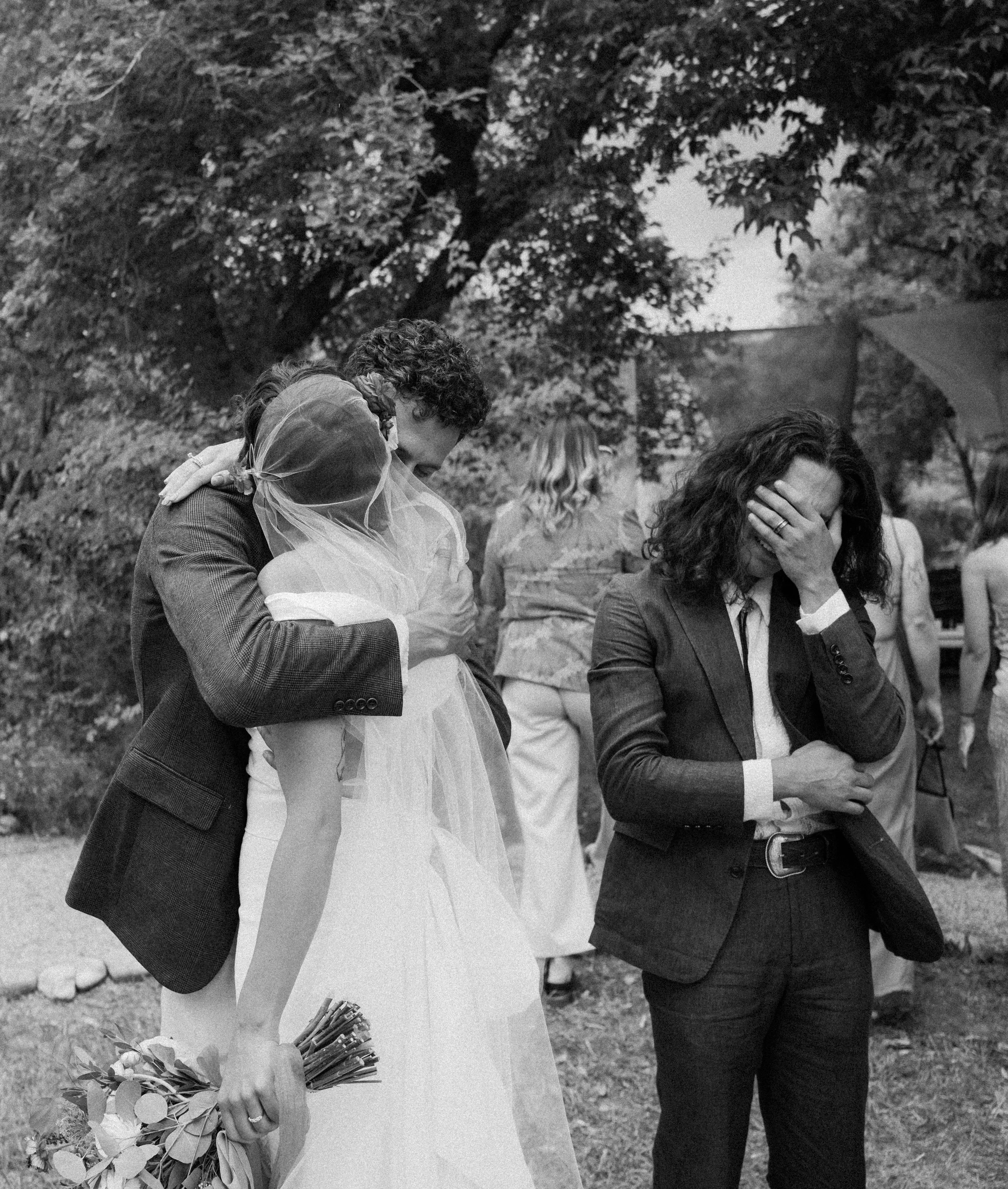 A black-and-white photo of a wedding scene outdoors. The bride and groom are embracing, with the bride holding a bouquet of flowers. A woman covers her face with her hand in front of them, standing among other guests.