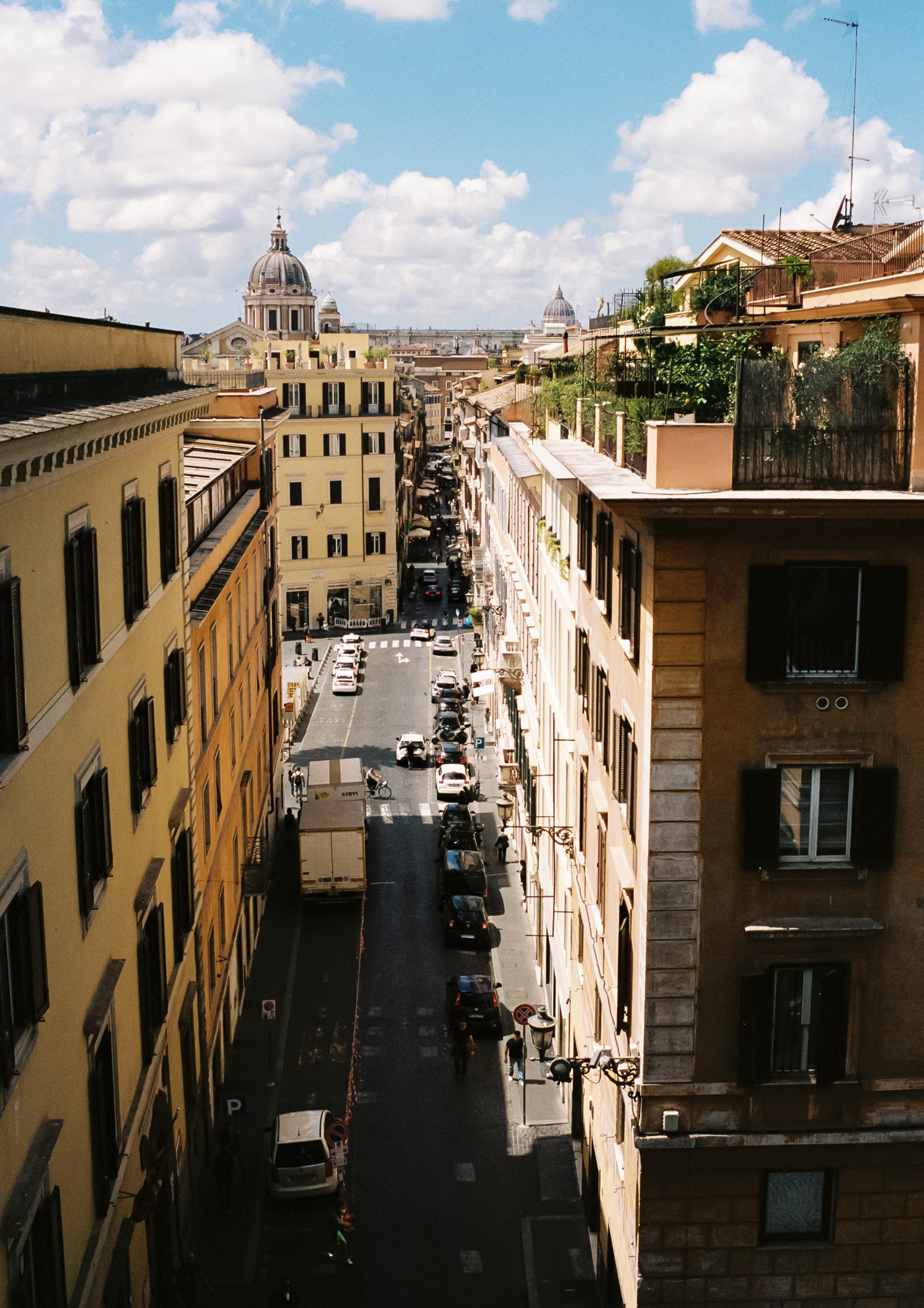 View of a narrow city street with parked cars, pedestrians, and multi-story buildings on both sides, leading toward domed rooftops in the distance under a partly cloudy sky.