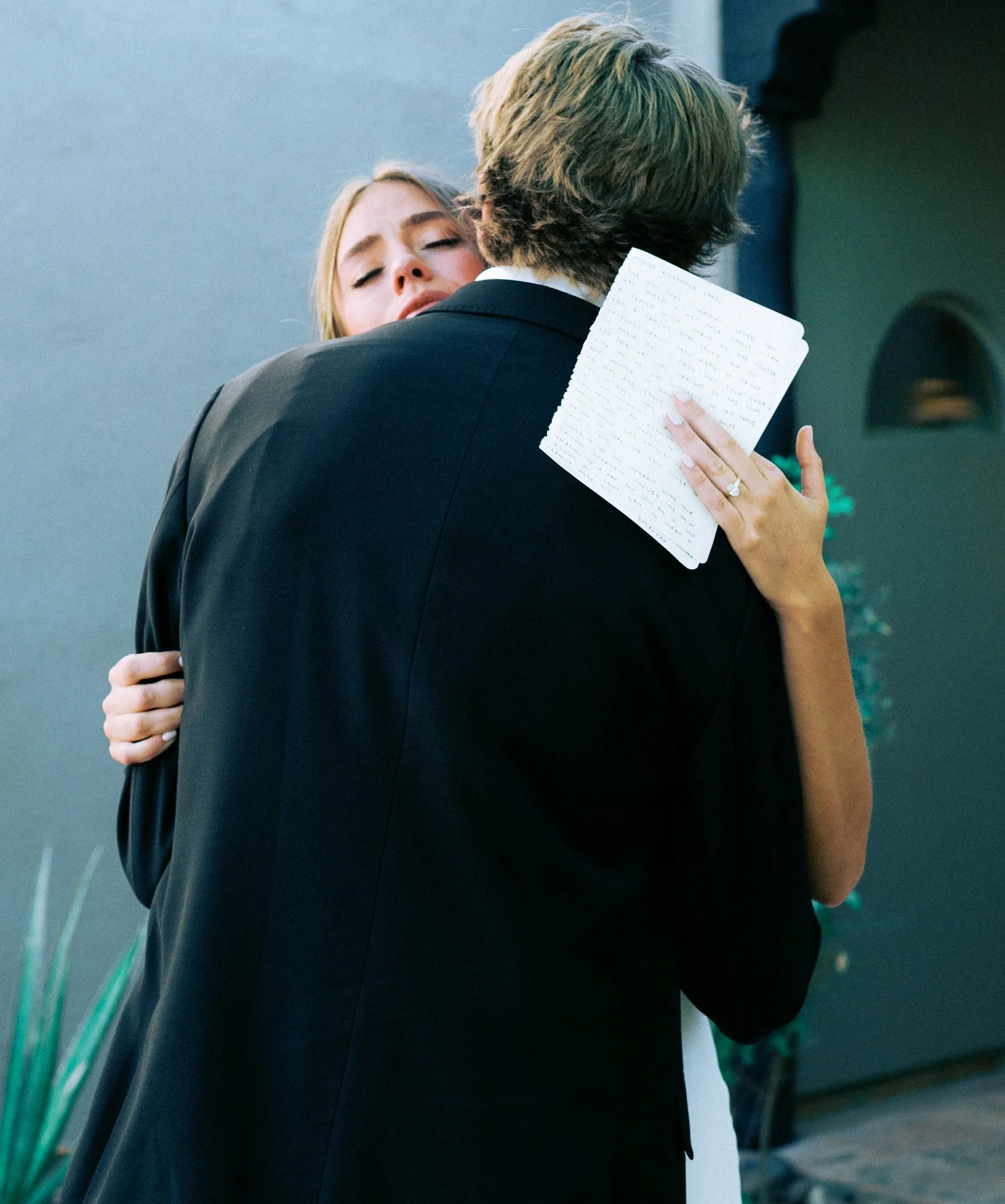 A woman with blonde hair and closed eyes embraces a man with brown hair, wearing a black suit, in an emotional hug. The woman is holding a handwritten letter and wearing a wedding ring. They are indoors with a gray wall and some potted plants visible in the background.