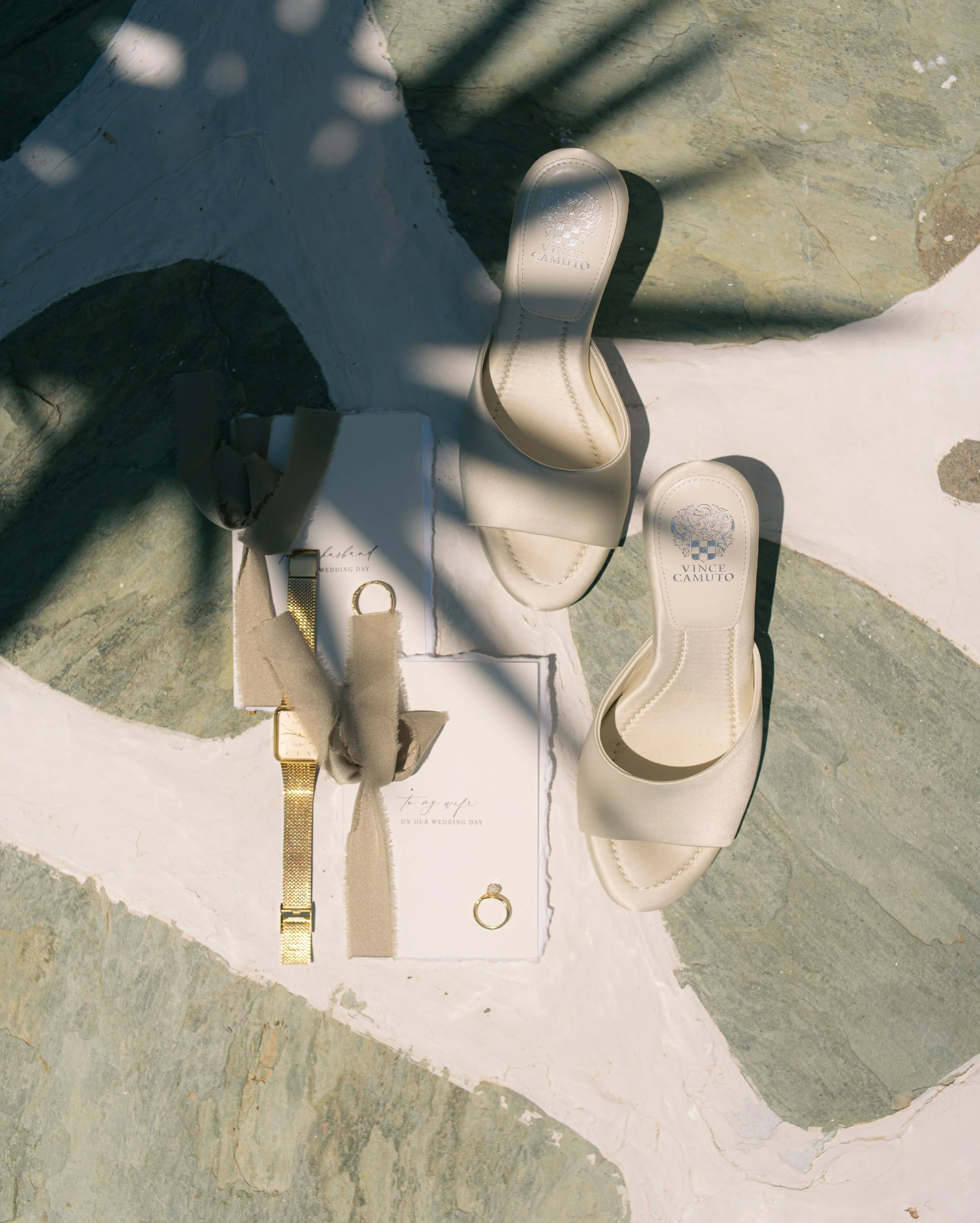 Cream-colored wedding shoes with Vince Camuto logo, gold jewelry including a ring and watch, and a wedding invitation on a textured stone surface with shadows.