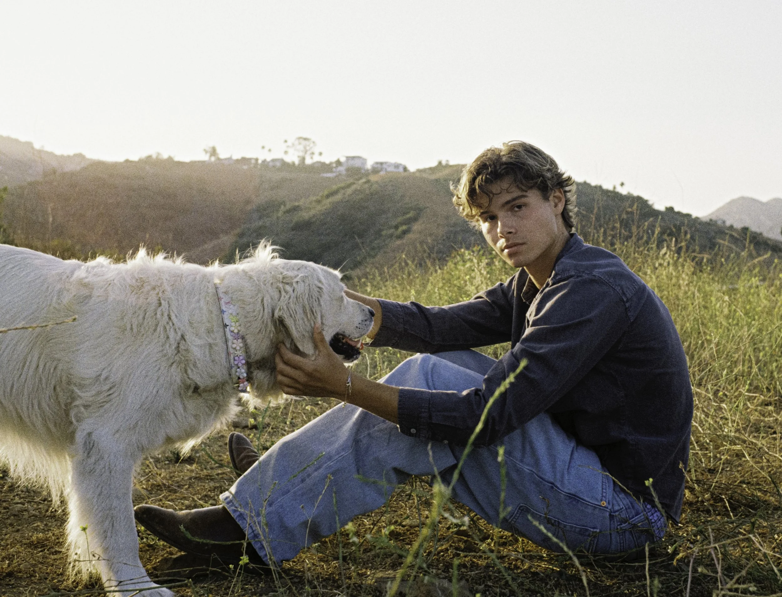 Young man sitting on grass with a golden retriever dog on a hillside with hills and houses in the background at sunset.