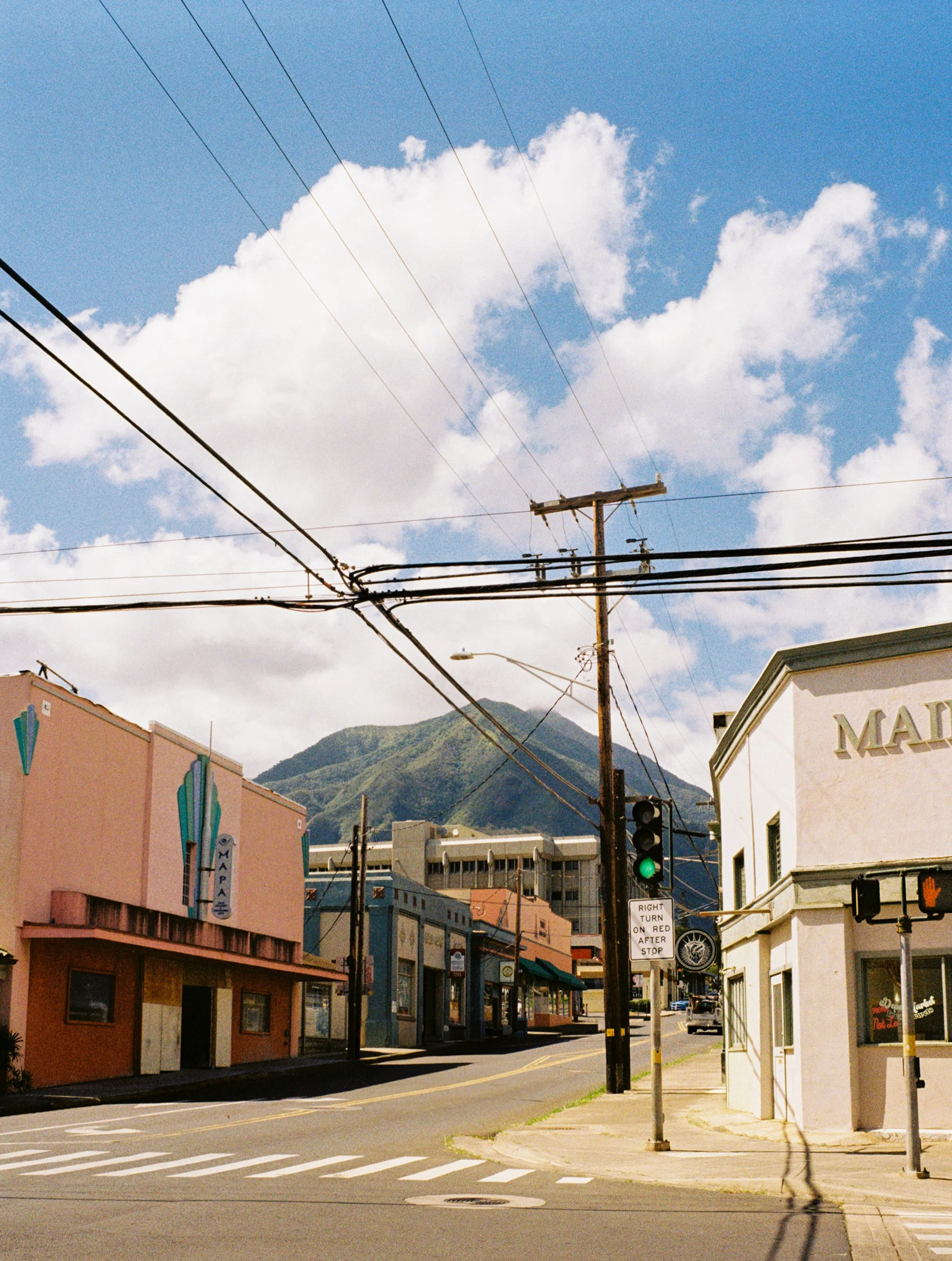 A street scene with colorful buildings, power lines crossing the sky, a mountain in the background, a traffic light showing green, and a sign indicating right turn on red after stop, under a partly cloudy blue sky.